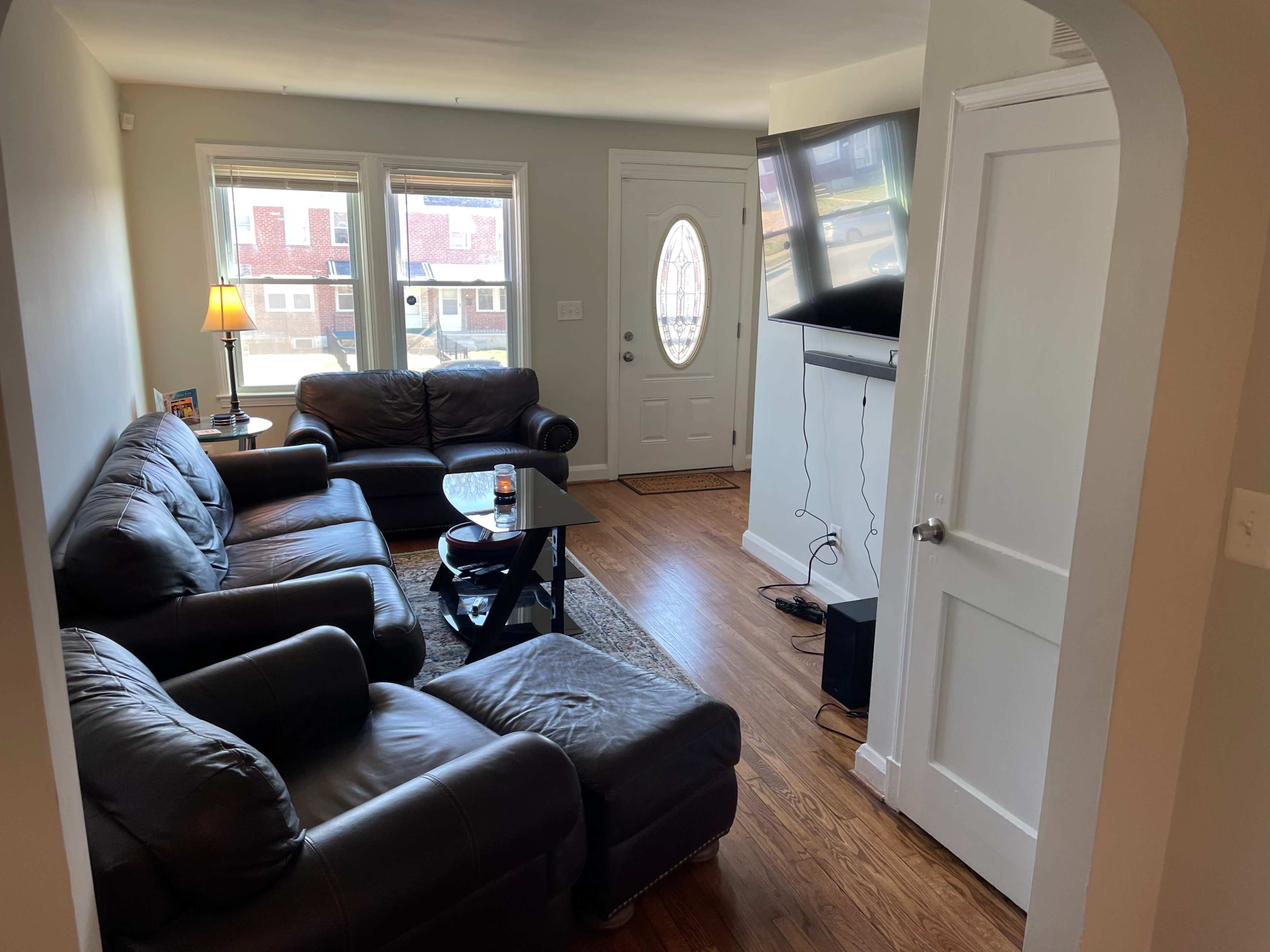 A cozy living room with two leather couches, a coffee table, and a television mounted on the wall, featuring large windows that allow natural light to enter.