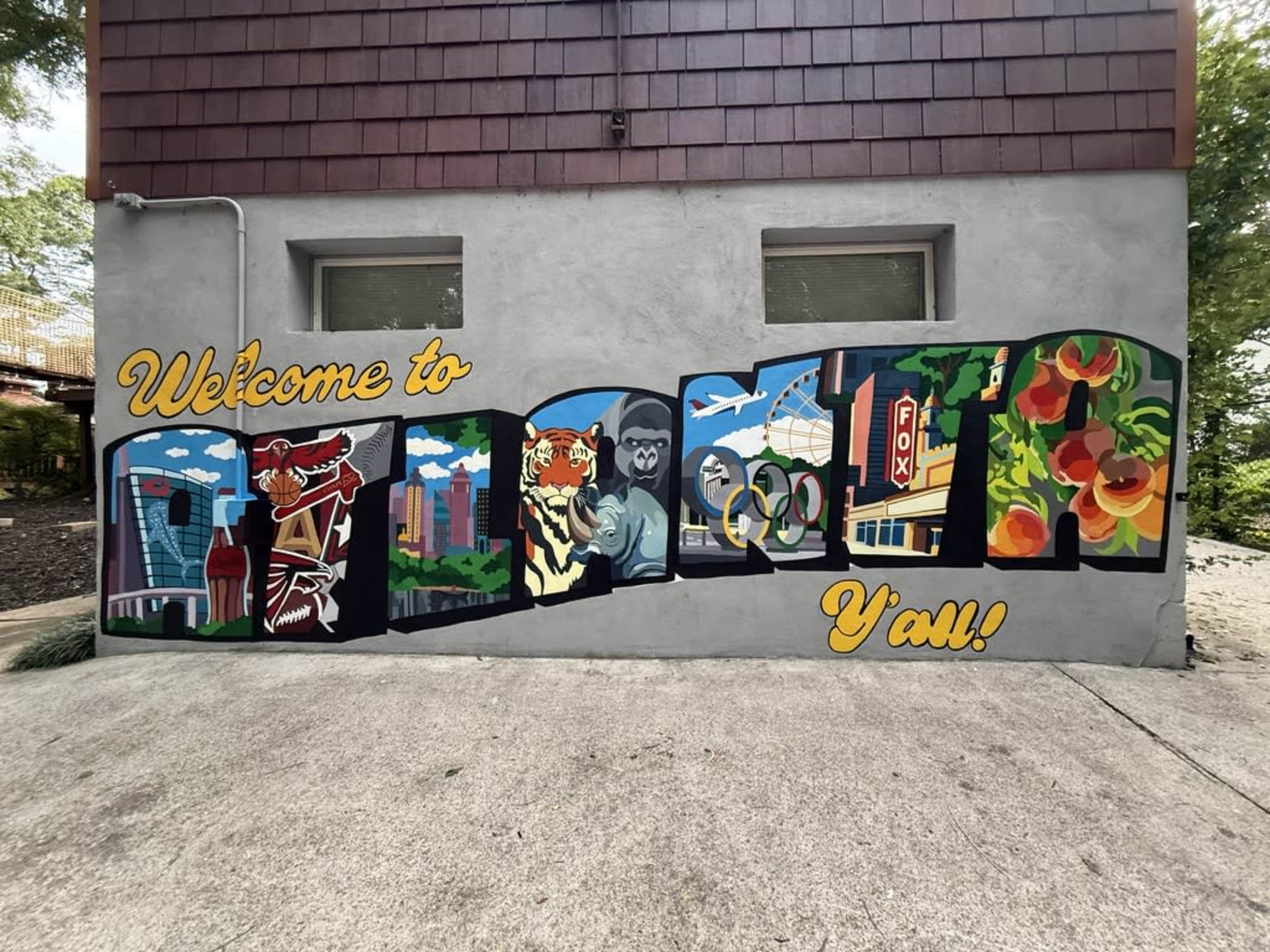 The image shows a colorful mural on a gray wall that reads "Welcome to [City's Name] You!" featuring various illustrations, including a skyline, a tiger, and a monkey.