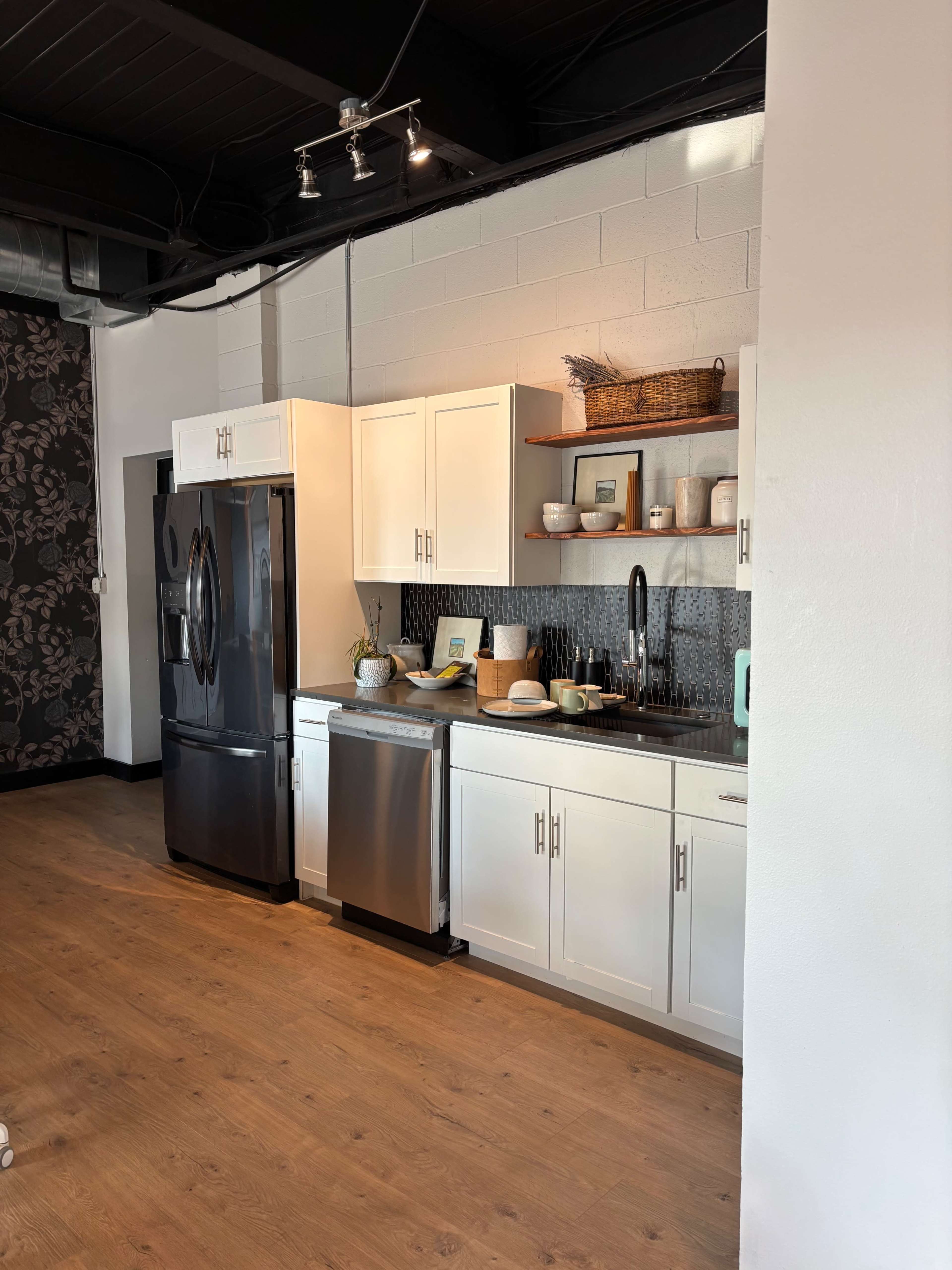 The image shows a modern kitchen with white cabinets, a black refrigerator, a stainless steel dishwasher, and wooden shelves displaying various kitchen items.