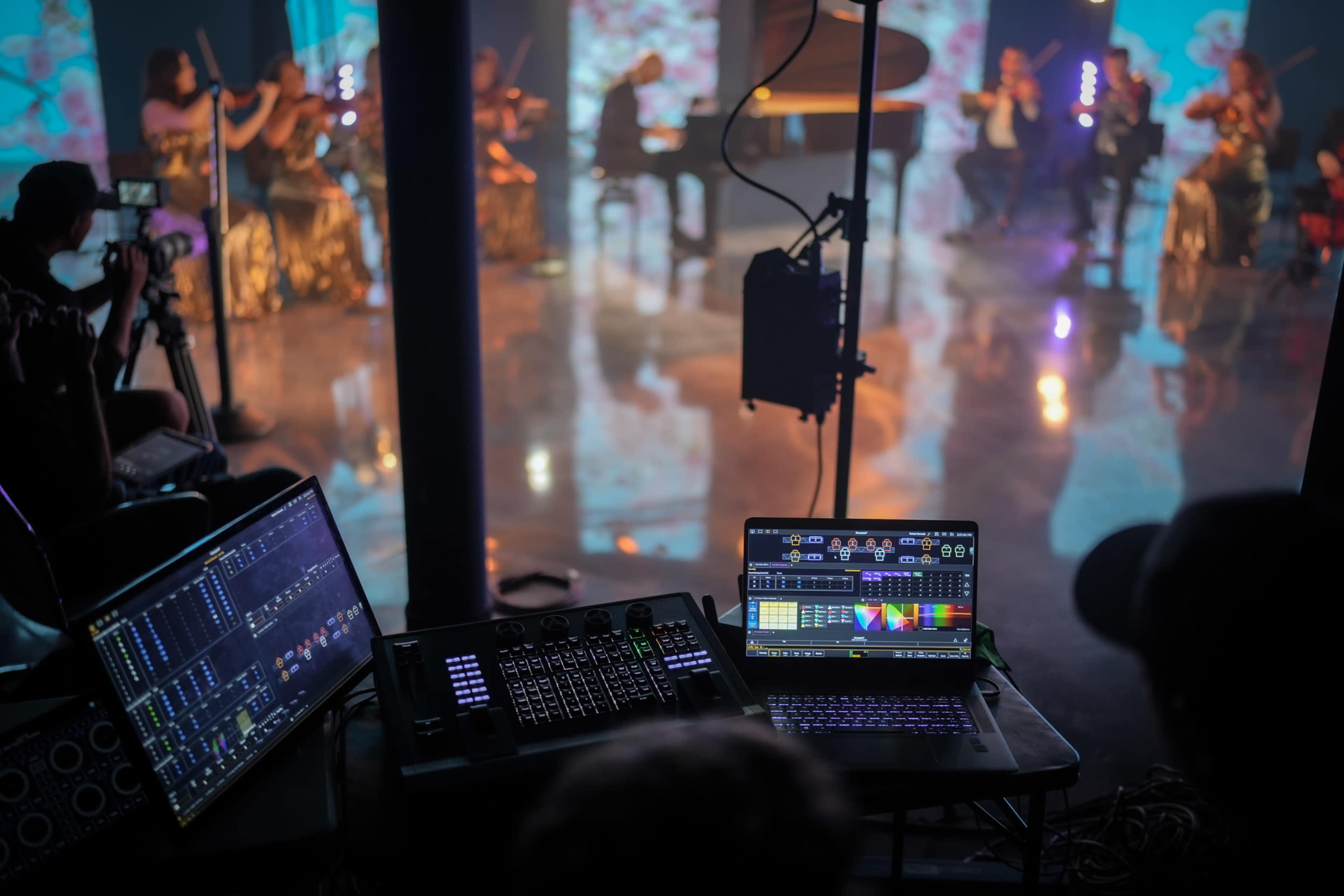 A live performance is taking place with musicians playing instruments on stage, while technical equipment displays lighting and sound controls in the foreground.