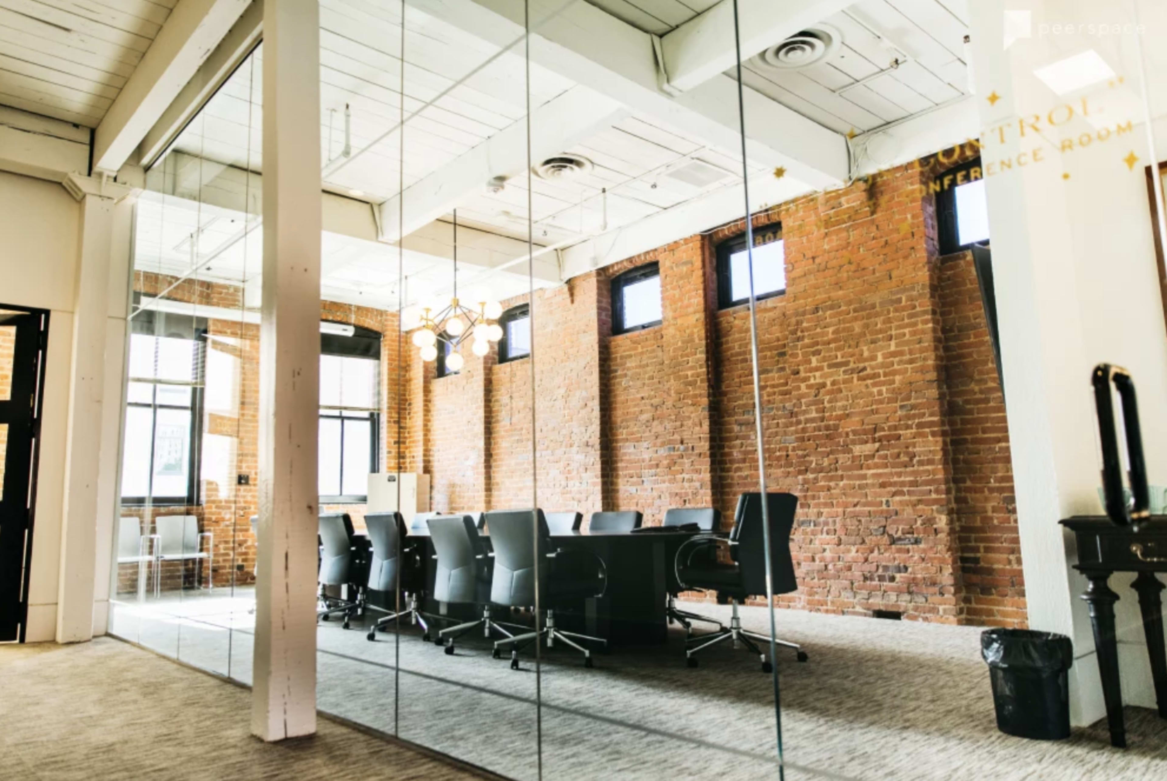 A modern conference room featuring a long table surrounded by black chairs, with large windows and exposed brick walls.