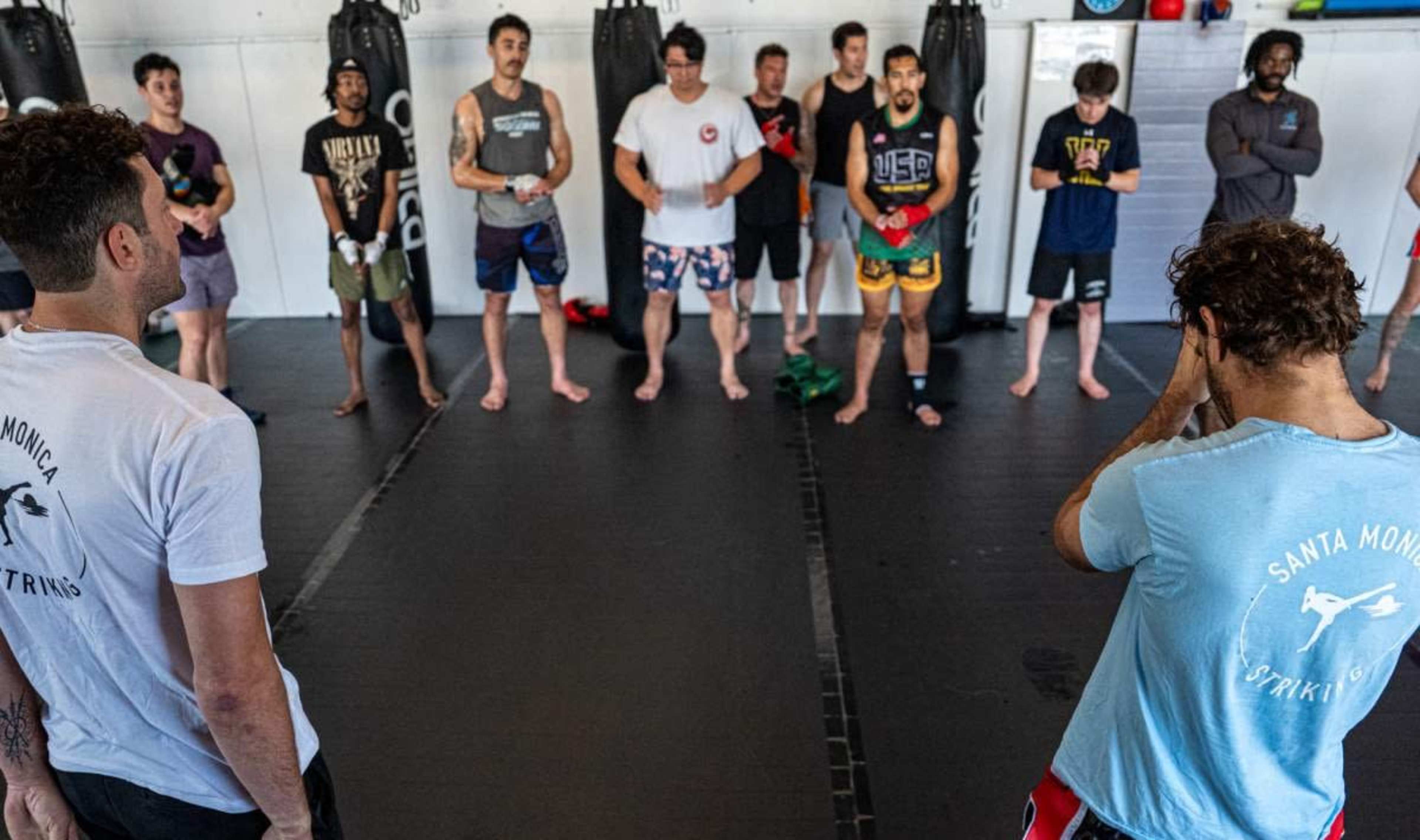 A group of men stands in a martial arts training facility, facing two instructors at the front.
