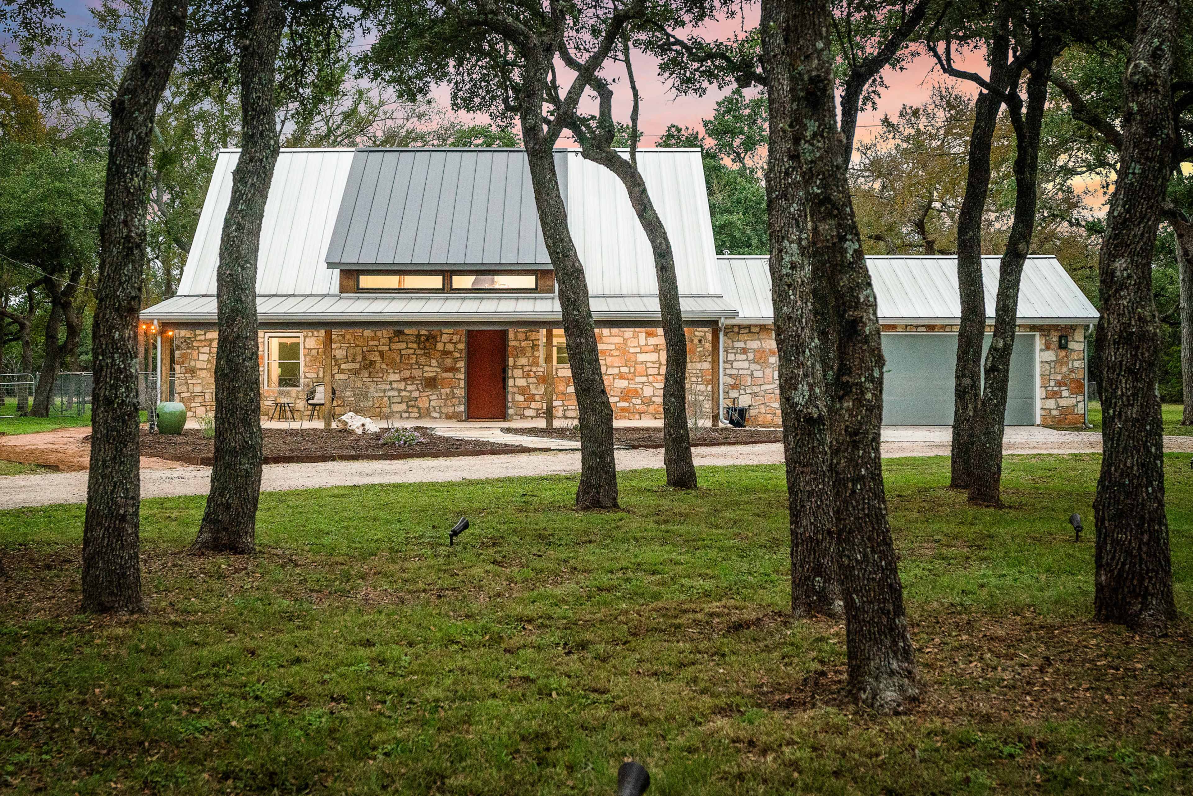 A modern stone house with a metal roof and a driveway is set among several trees at dusk.