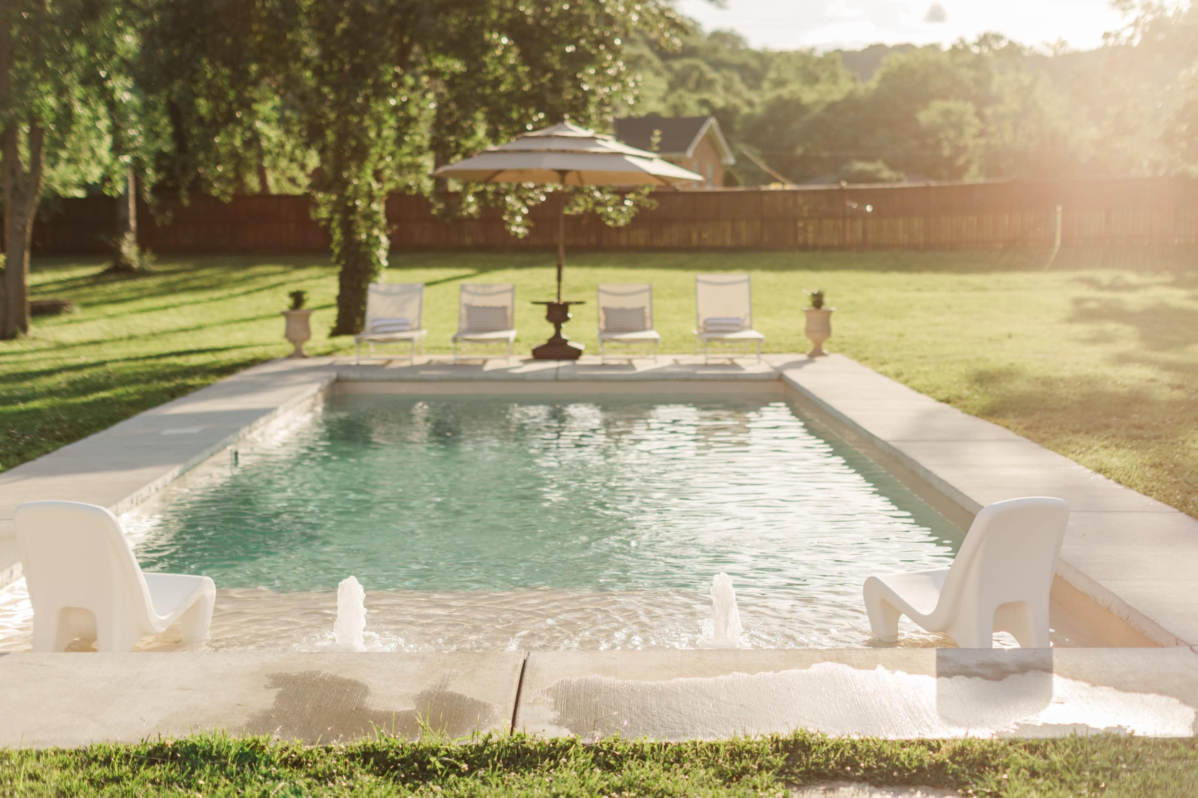 A clear swimming pool is surrounded by a grassy area and several white lounge chairs under an umbrella.