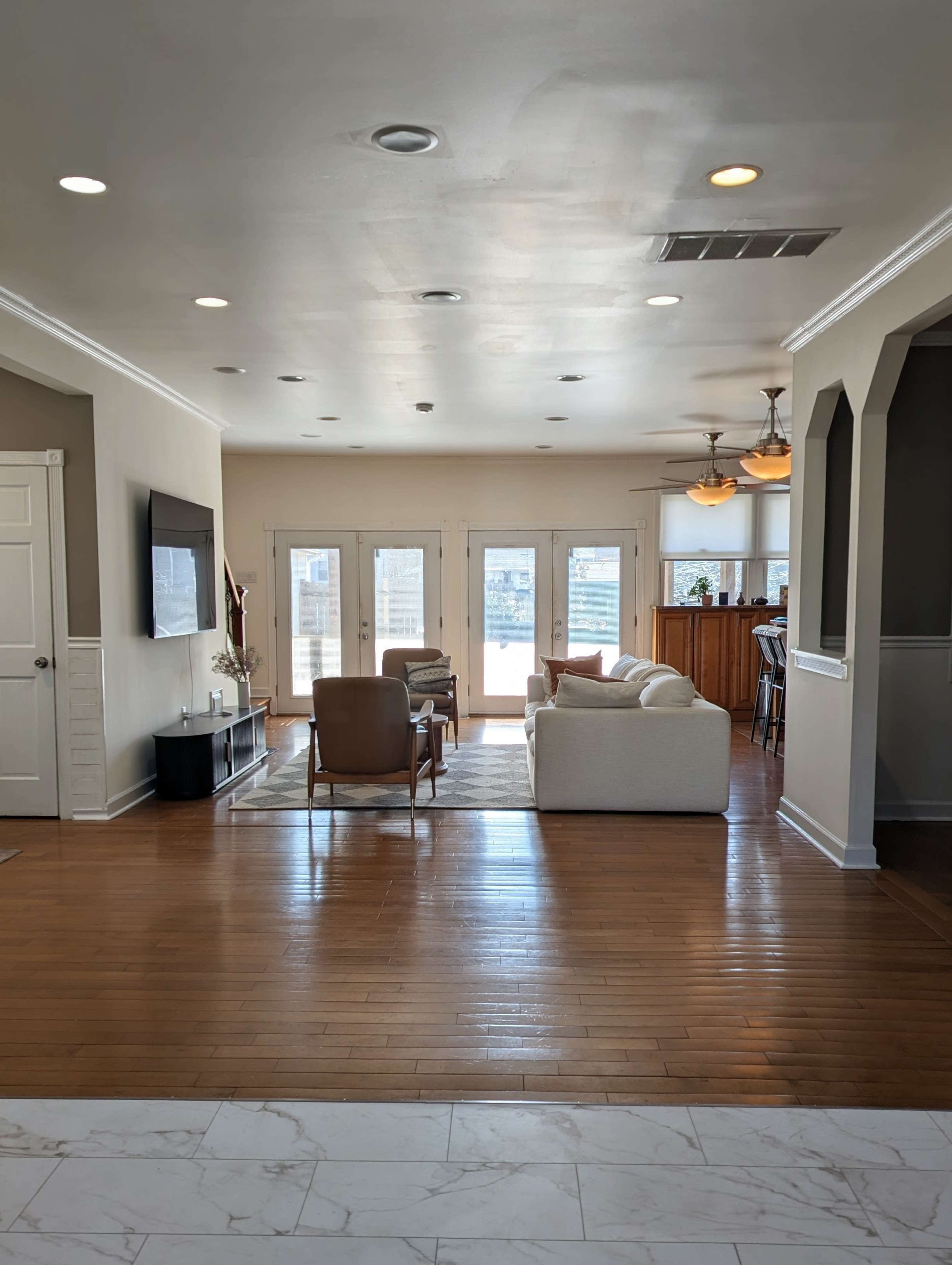 The image shows a spacious living room with hardwood floors, a white sofa, two armchairs, and large windows that lead to an outdoor area.