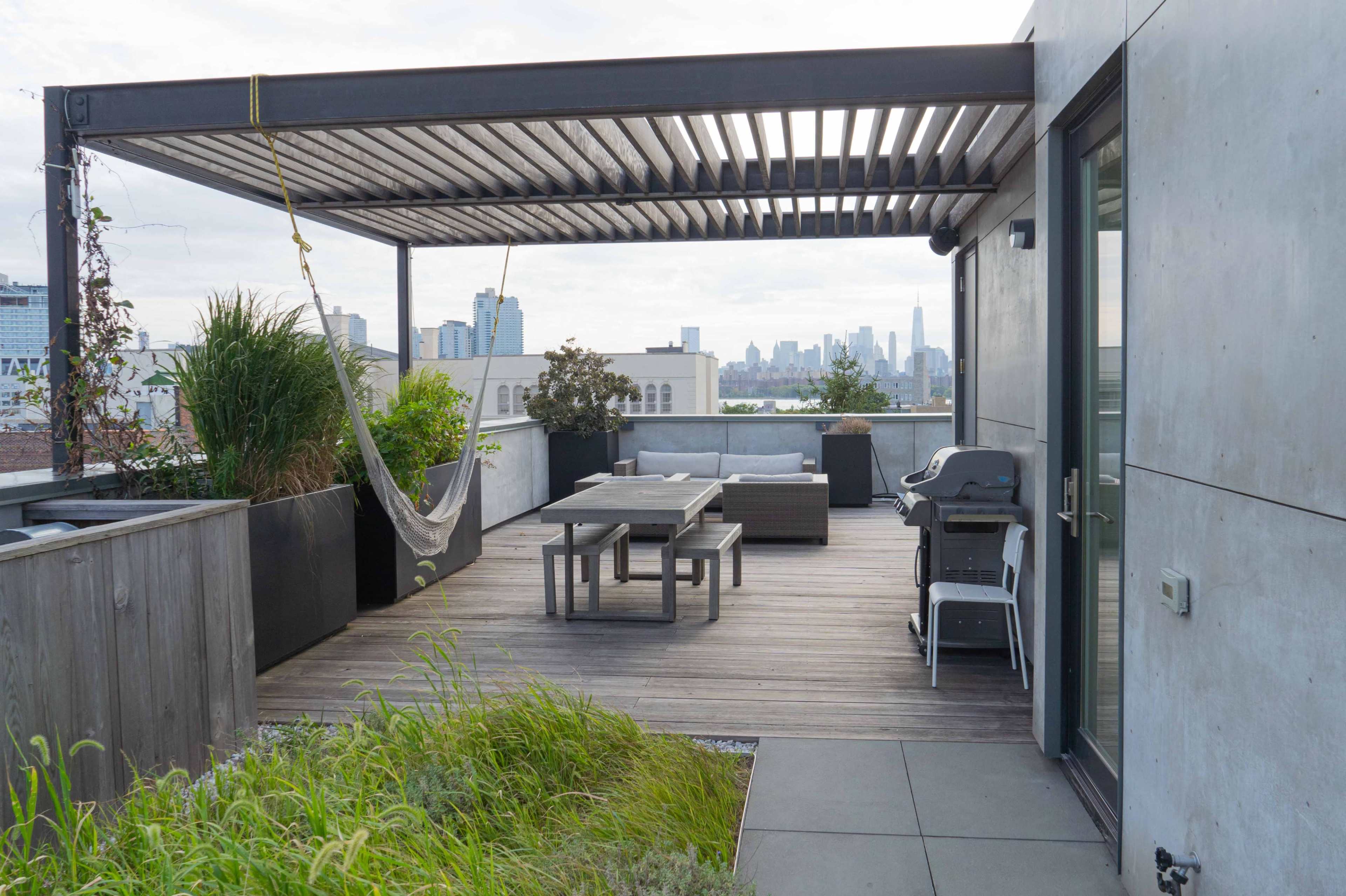 The image shows a modern rooftop terrace with a wooden deck, a dining table, a hammock, potted plants, and a city skyline in the background.