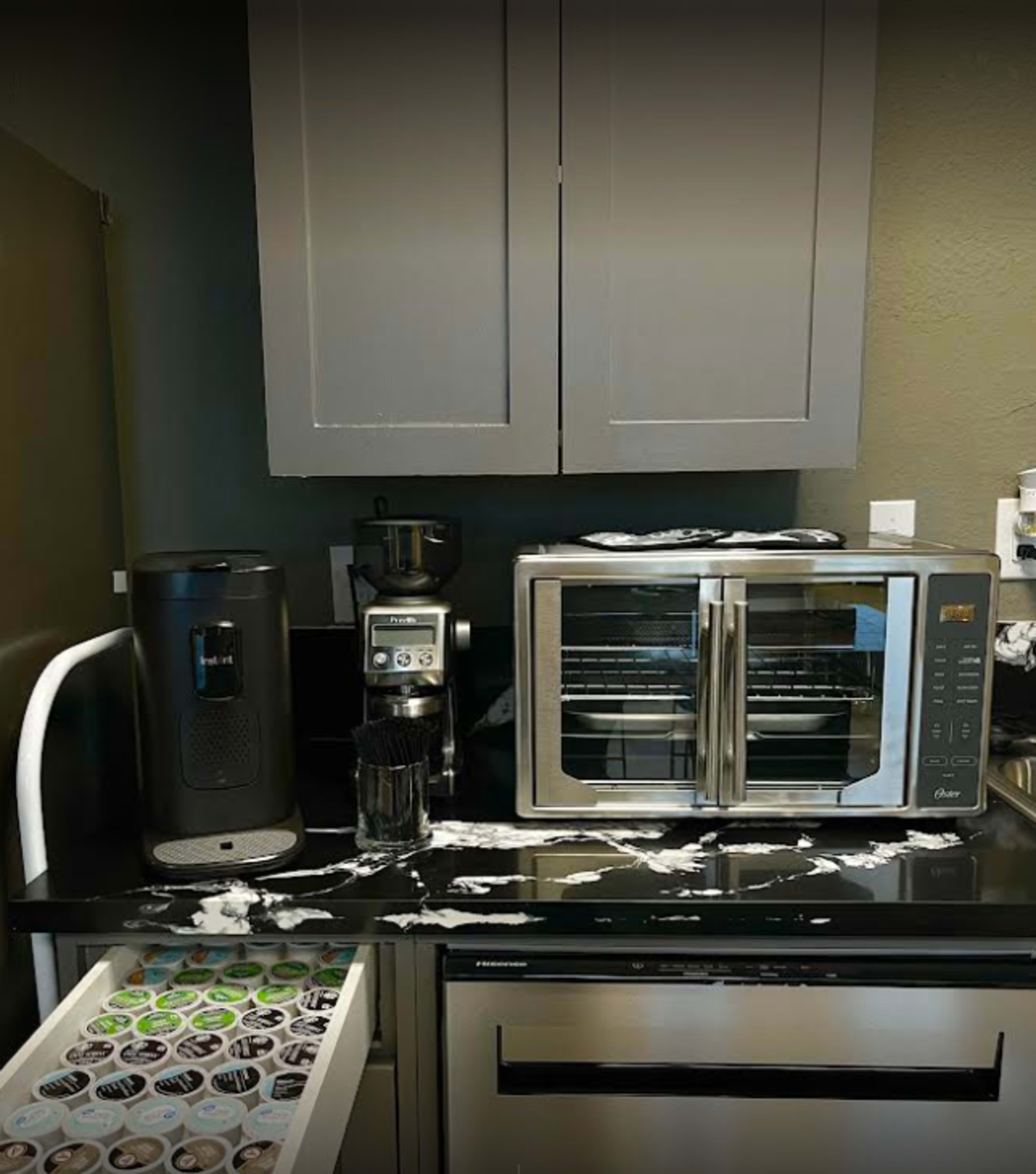A kitchen countertop features a coffee maker, a toaster oven, and a drawer filled with coffee pods, with what appears to be spilled liquid across the surface.
