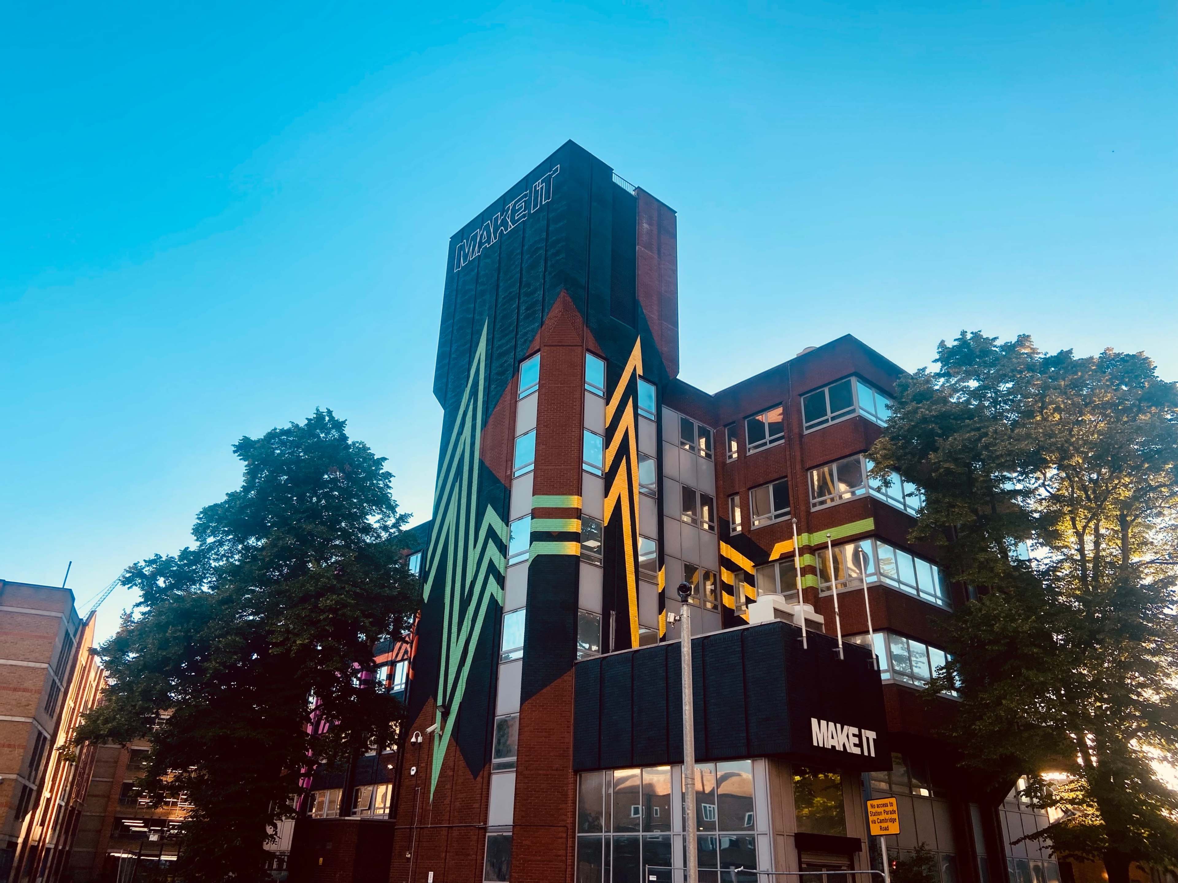 A multicolored modern building with a tower featuring the words "MAKE IT" is surrounded by trees and a clear blue sky.