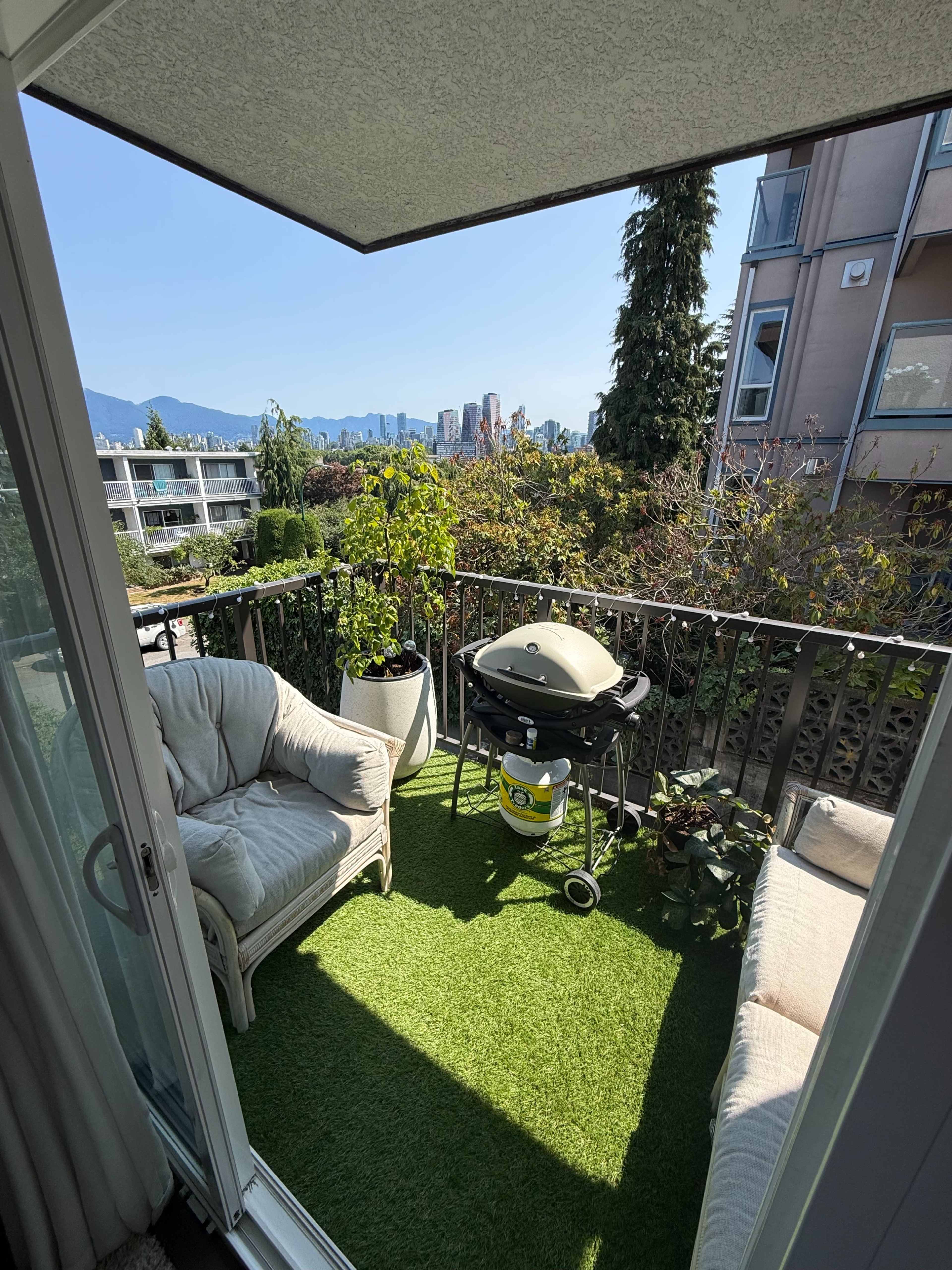 A small balcony features two chairs, a barbecue grill, potted plants, and a view of the city skyline and mountains in the background.