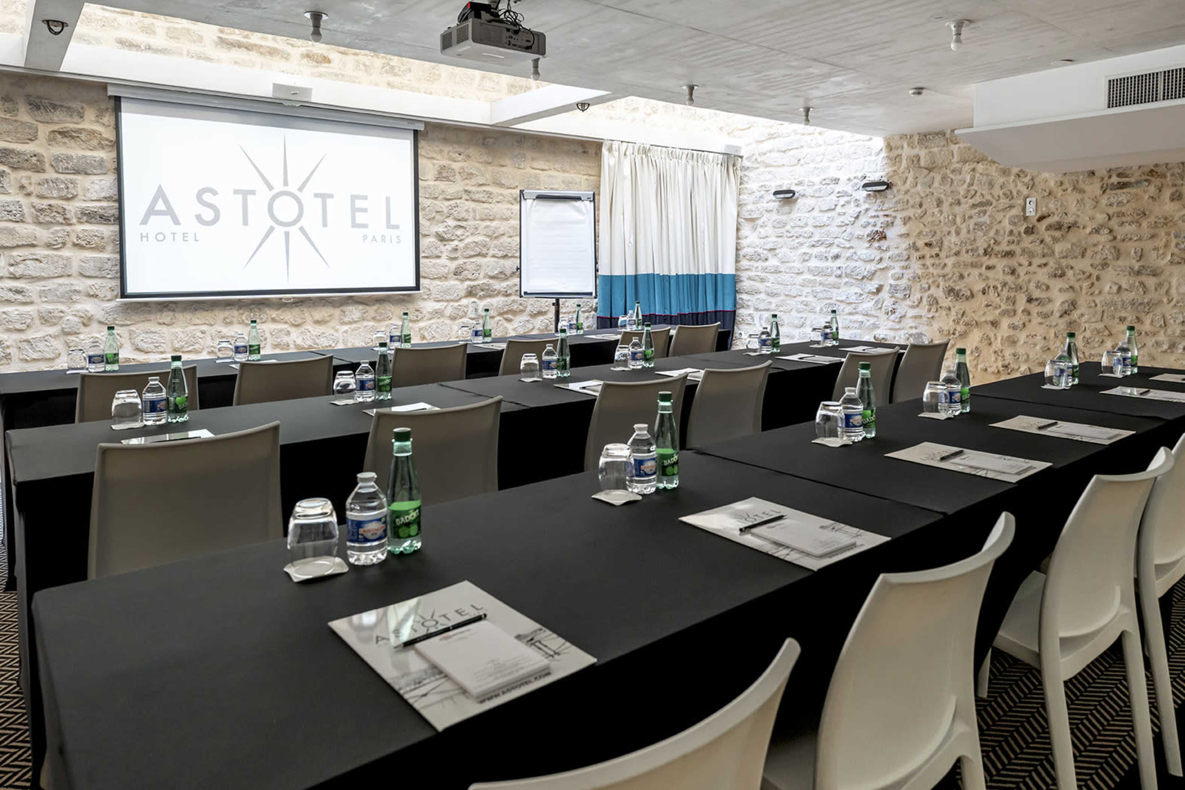 A conference room setup in the Astotel Hotel in Paris features rows of tables with water bottles and notepads, facing a large screen displaying the hotel logo.