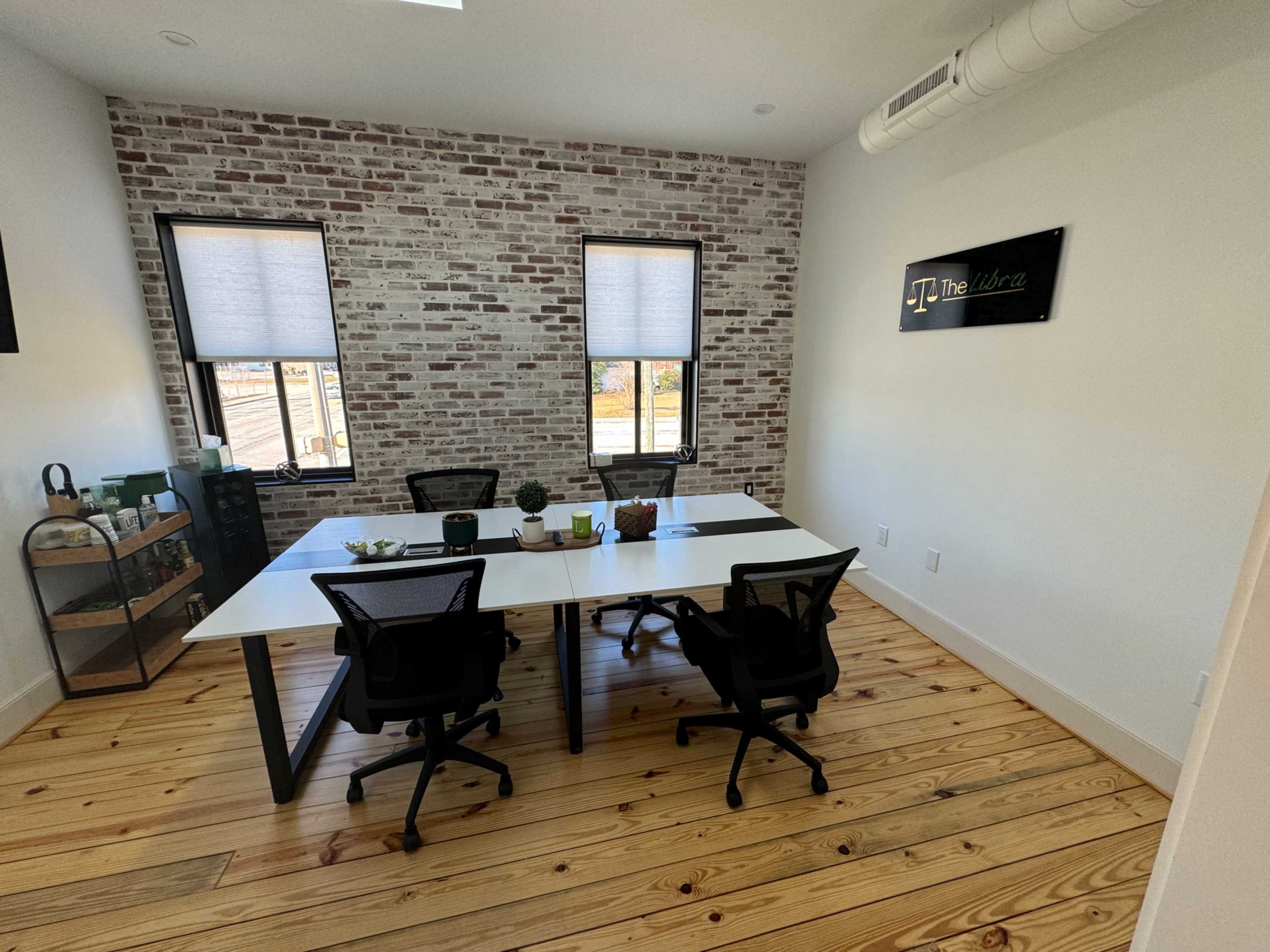 A modern office features a white table surrounded by black chairs, with exposed brick walls and large windows.