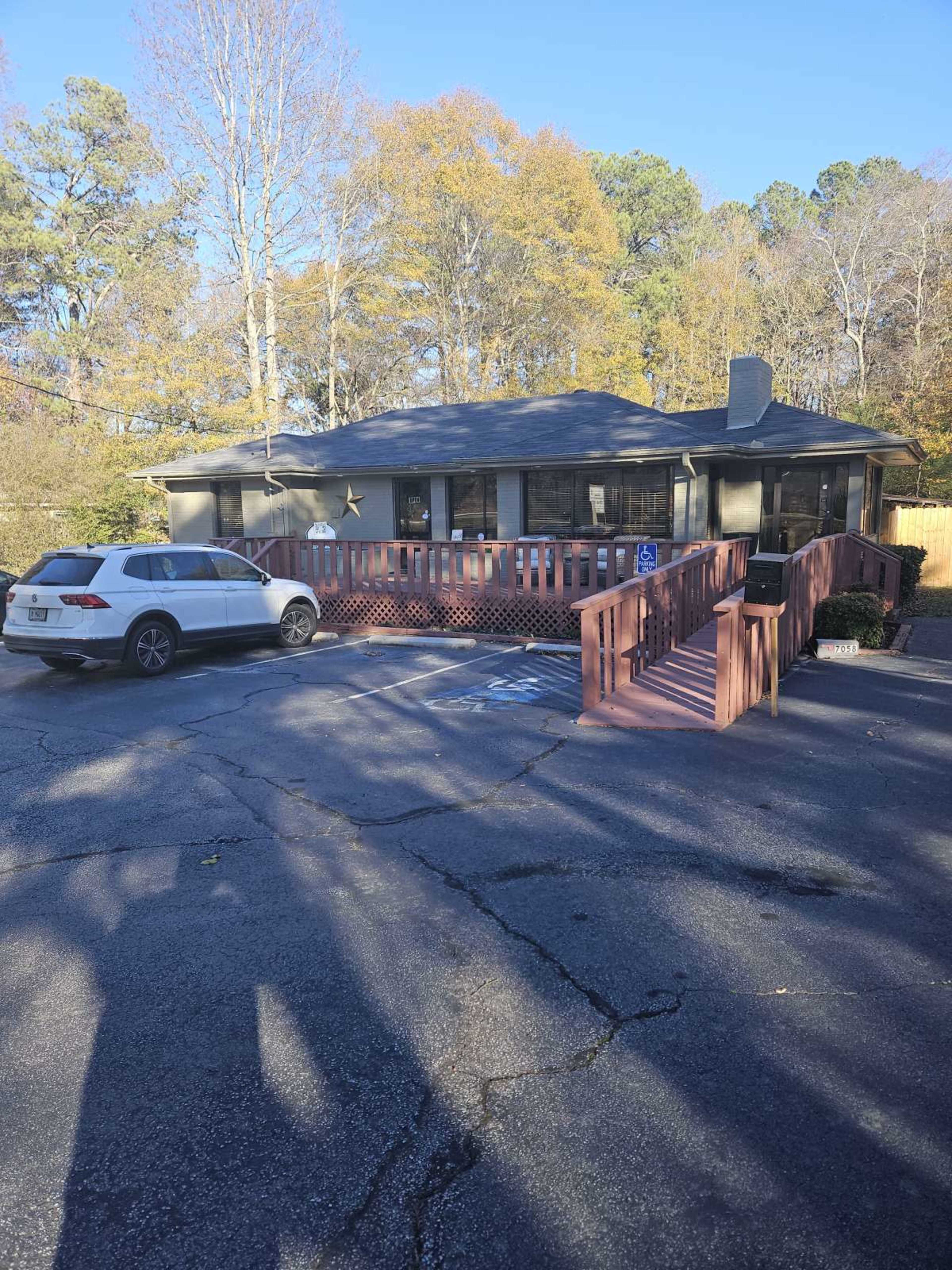 The image shows a single-story house with a wooden ramp leading to a front porch, parked car in the driveway, and surrounding trees in the background.