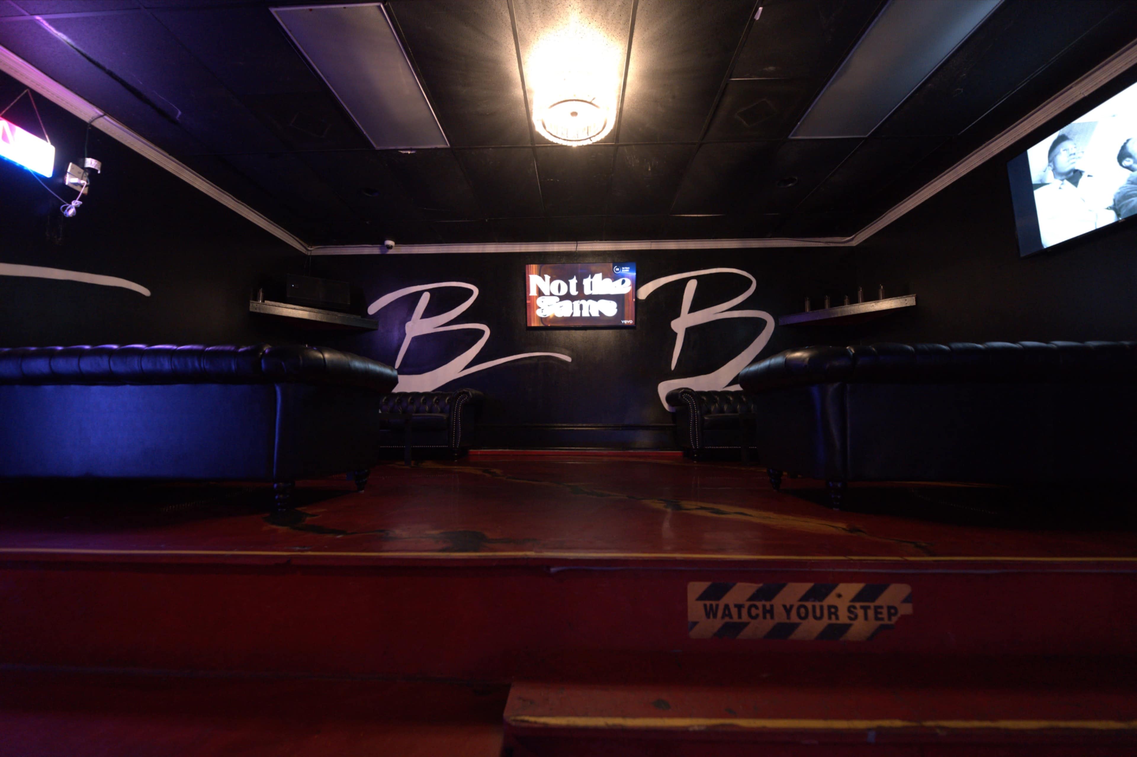 The image shows a dimly lit lounge area with black leather couches, a neon sign that reads "Not the Same," and a warning sign on the floor that says "WATCH YOUR STEP."