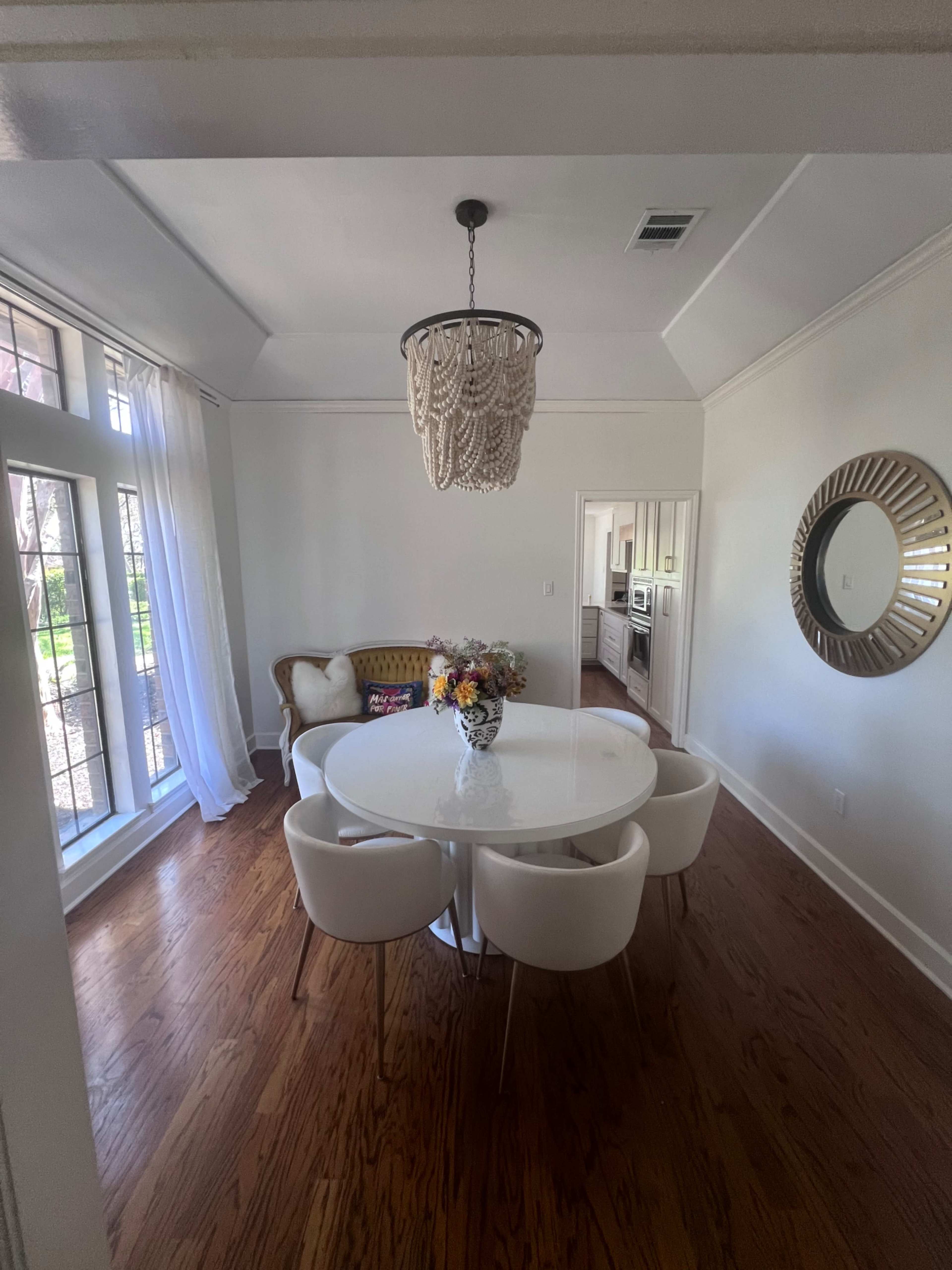 A dining area features a round table surrounded by four white chairs, with a chandelier overhead and large windows letting in natural light.
