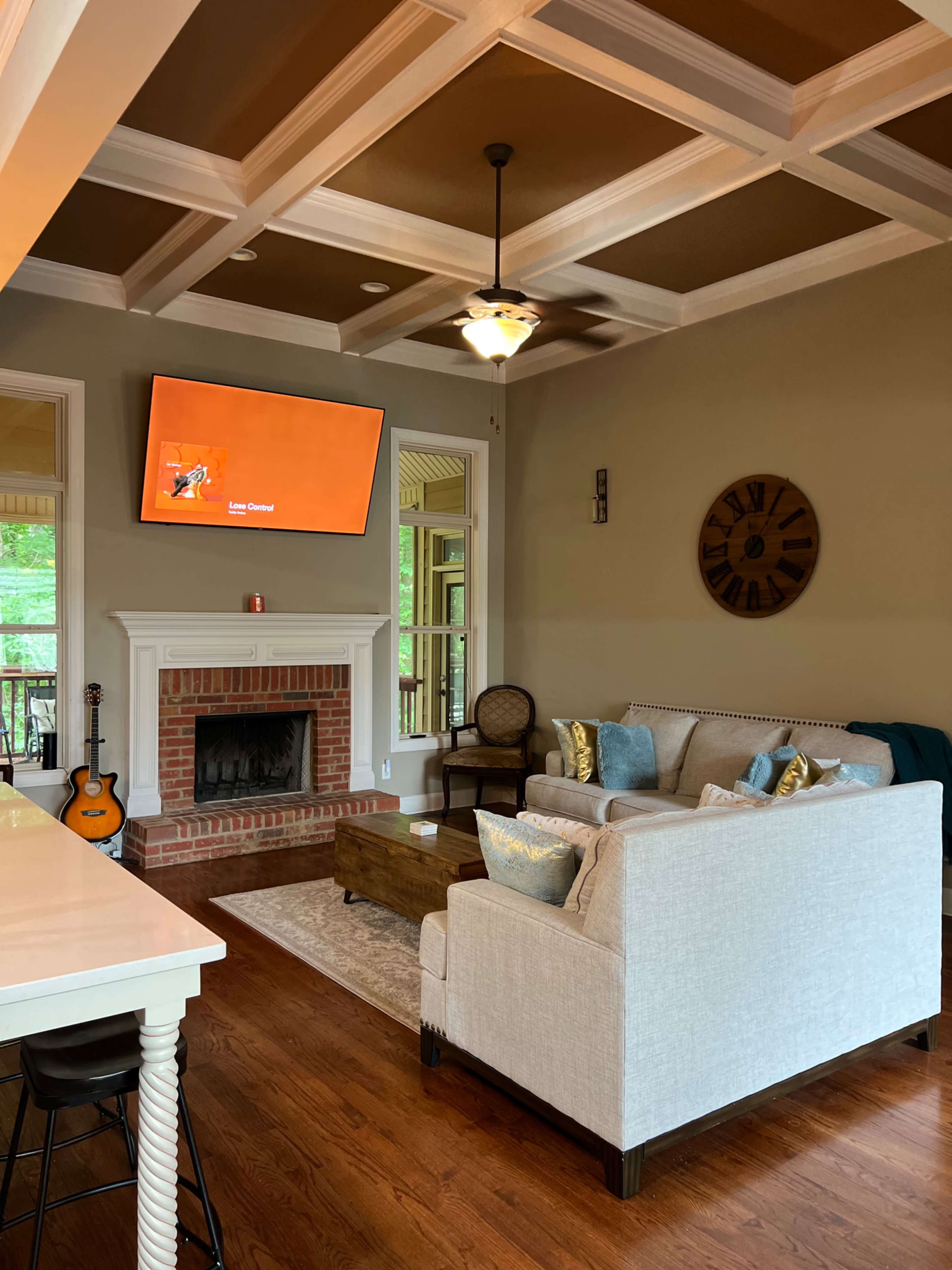 The living room features a sofa with decorative pillows, a coffee table in front, a fireplace, and a mounted television, all illuminated by natural light through large windows.