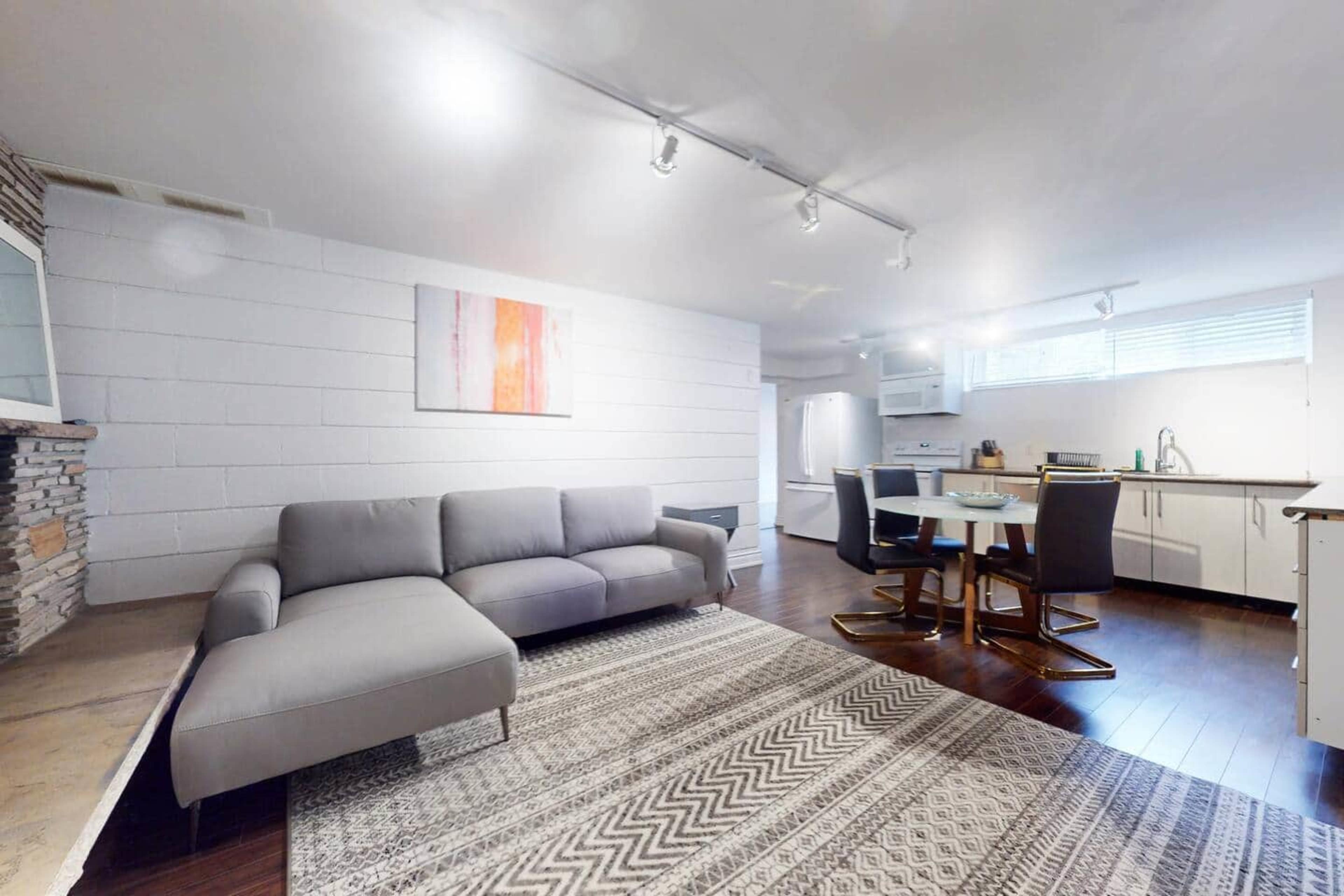 The image shows a modern living space featuring a gray sectional sofa, a round dining table with four chairs, and a kitchen area with white cabinetry.