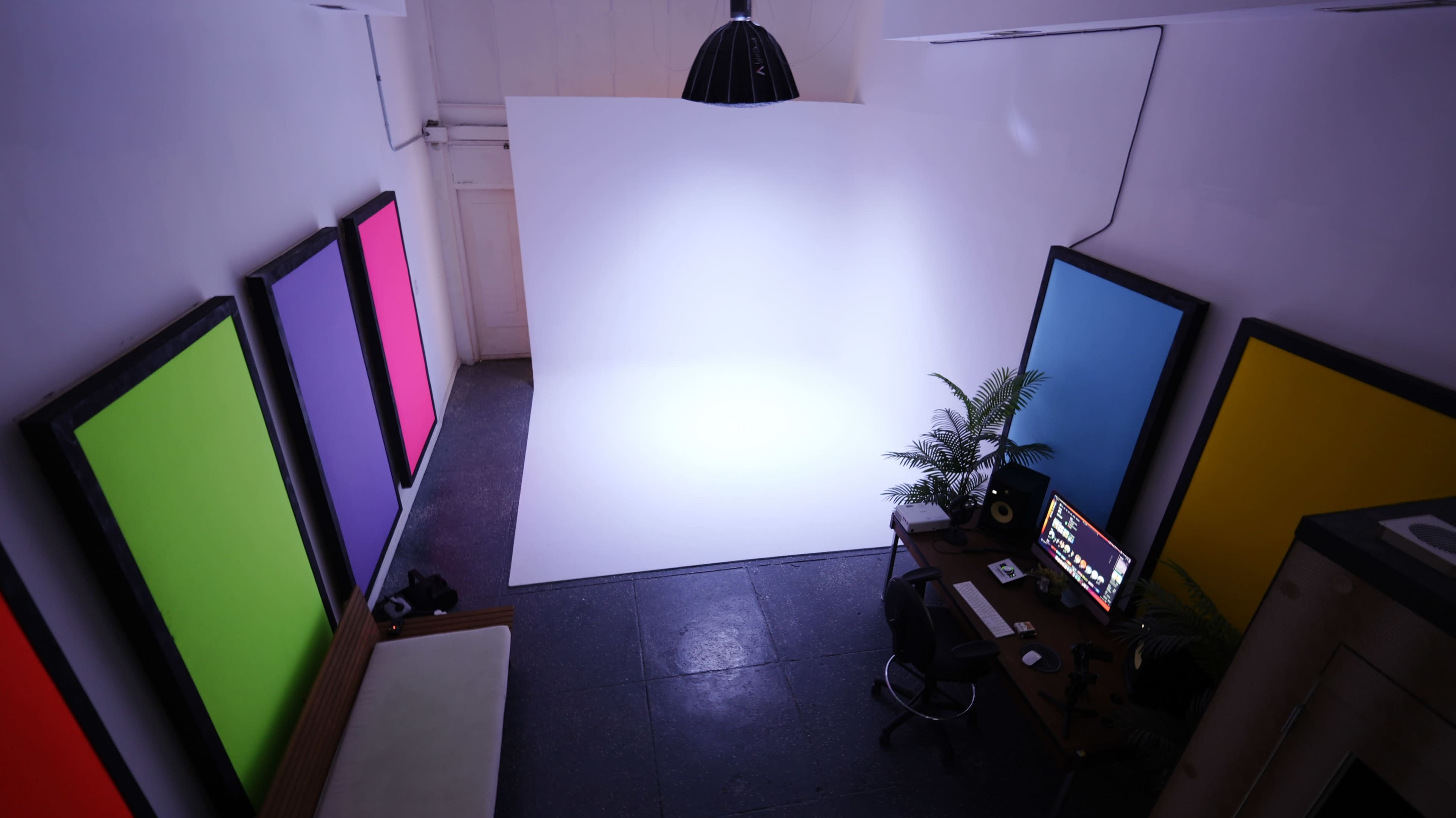The image shows a small studio space with a white backdrop, a desk with computer equipment, and colorful screens on the walls.