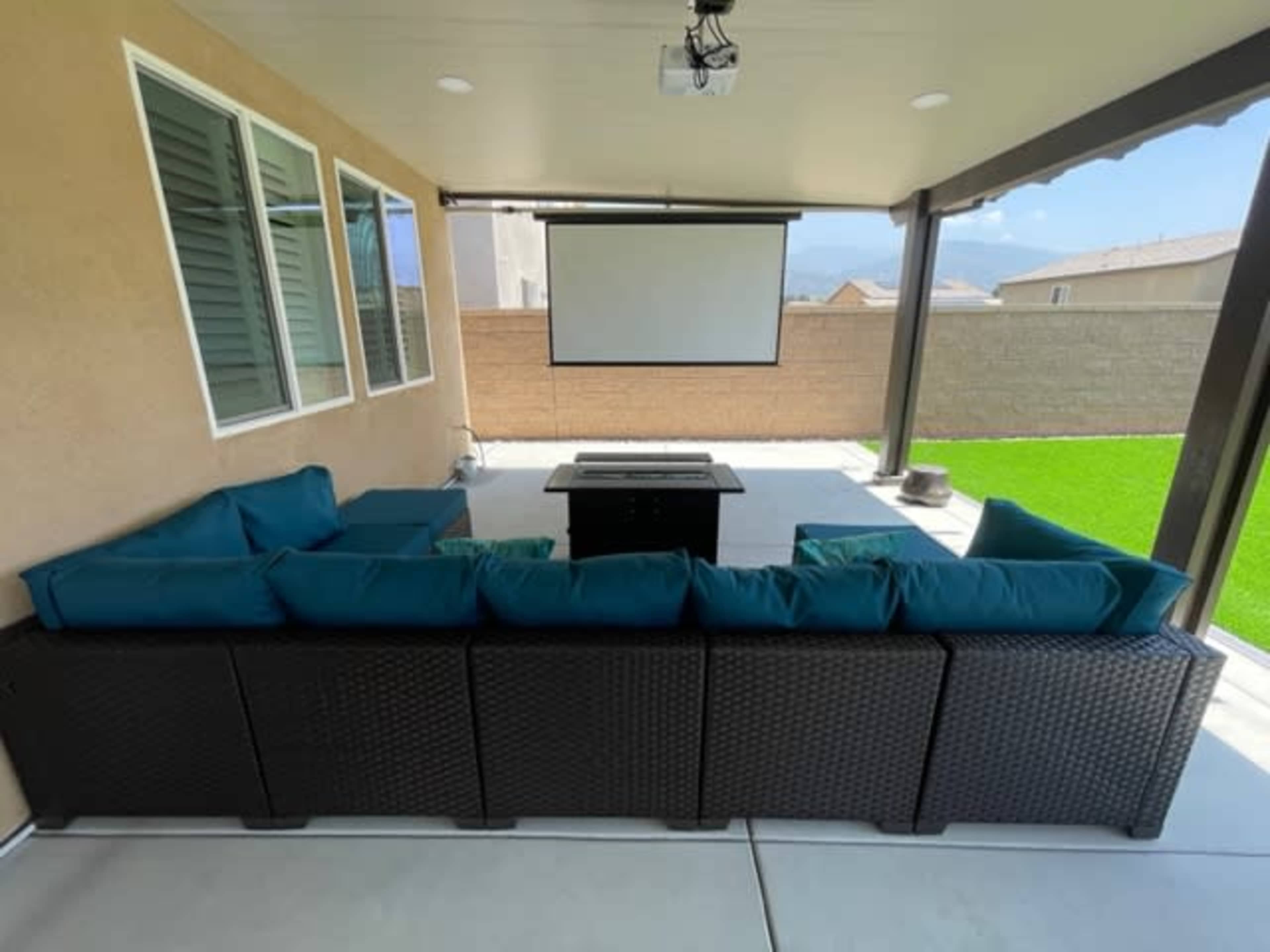 A covered outdoor patio features a sectional sofa with teal cushions facing a projector screen on the wall.