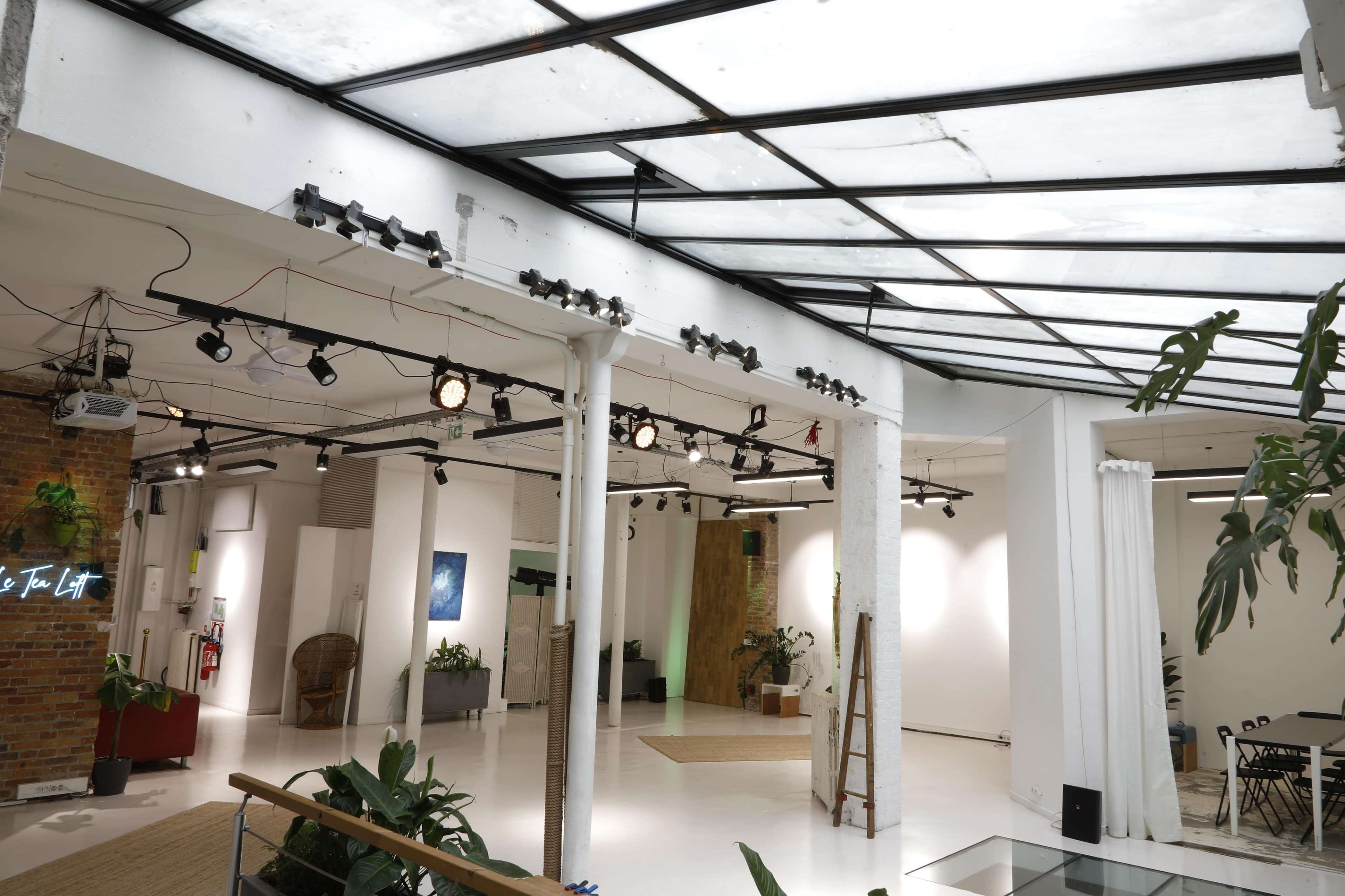 The image shows a spacious, well-lit interior featuring a mixture of white walls and exposed brick, with industrial lighting and indoor plants.