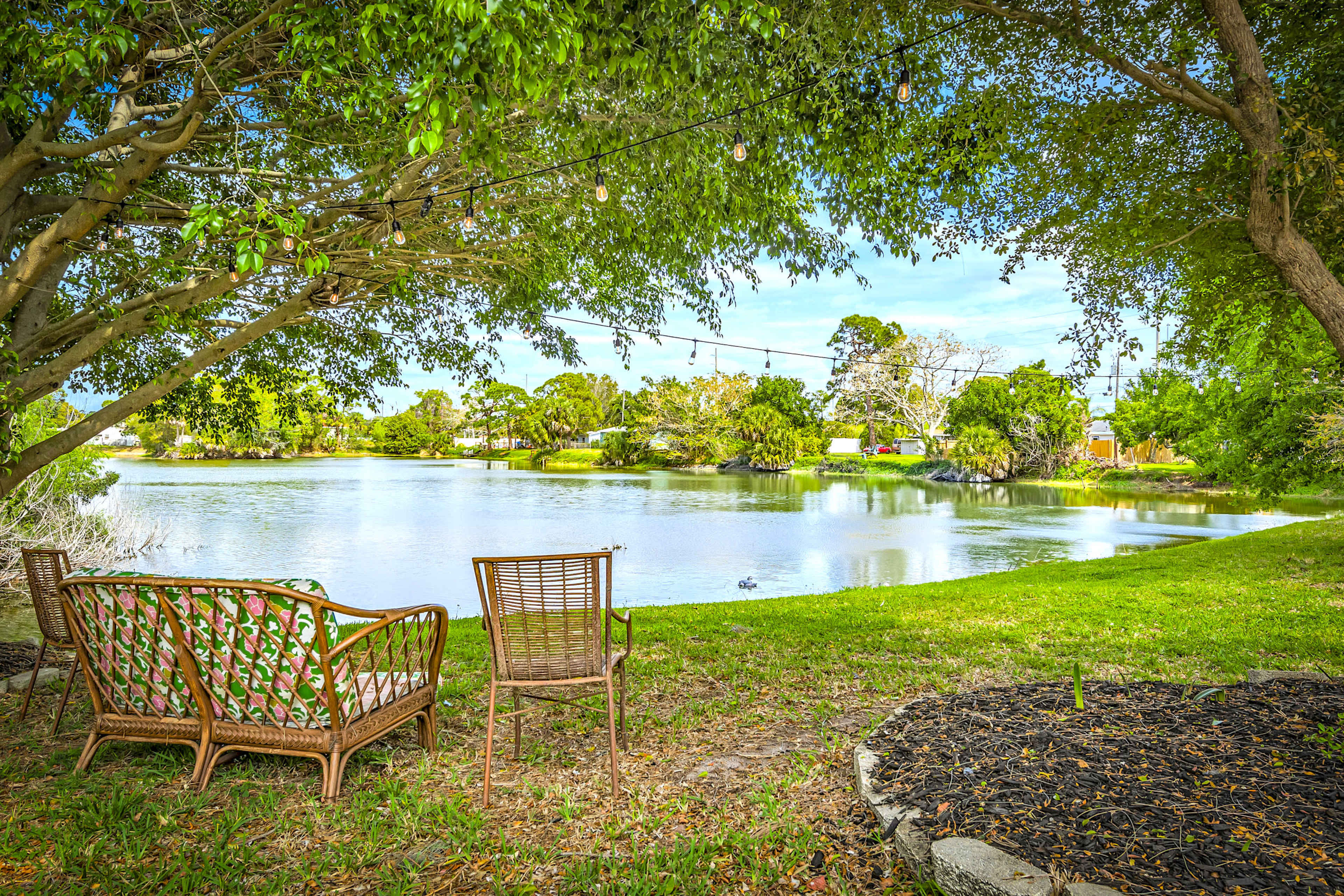 A pair of wicker chairs is positioned near a calm lake surrounded by trees and greenery, with decorative string lights overhead.