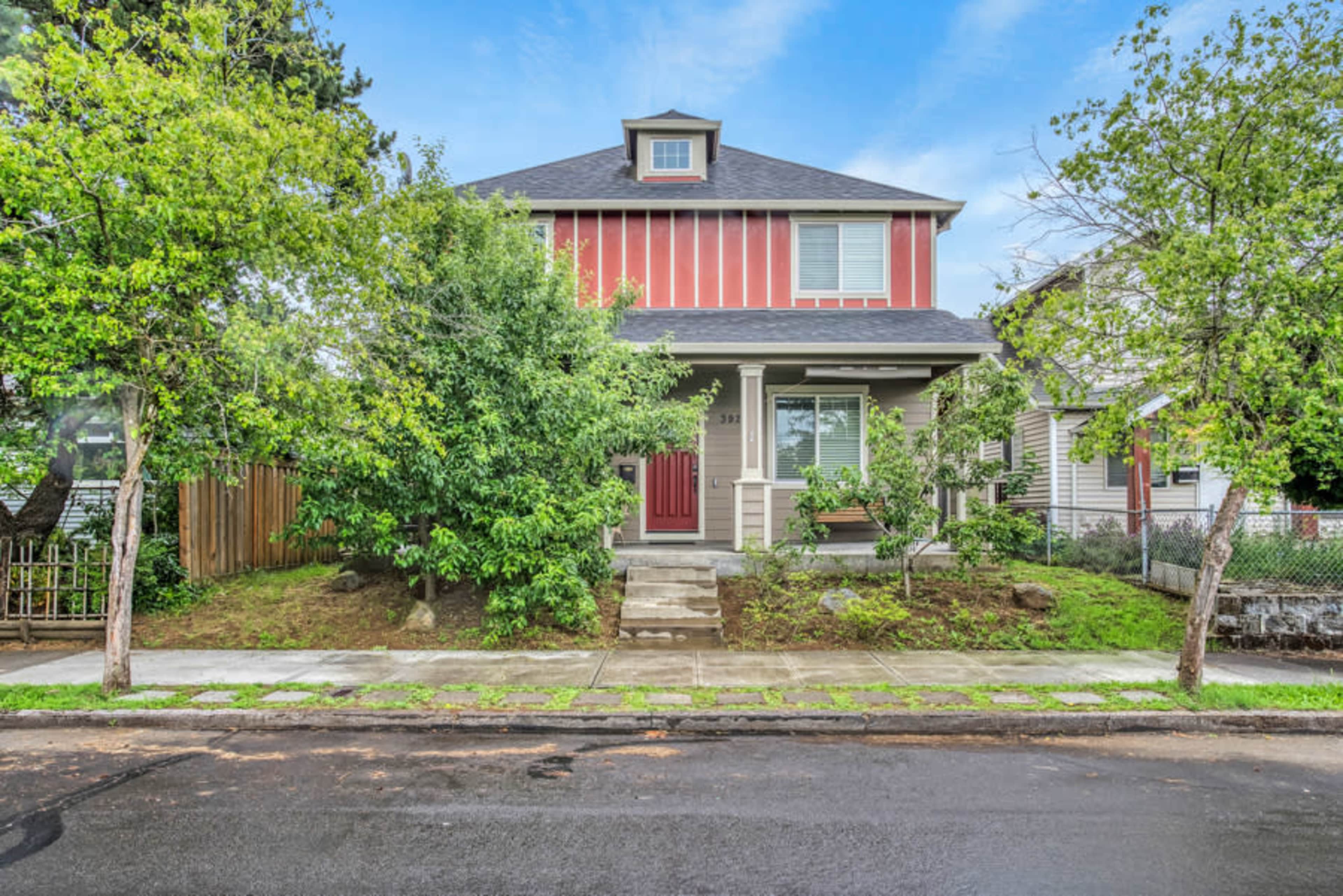 A two-story house with red and beige siding, a gabled roof, and a front porch is surrounded by trees and shrubs on a residential street.