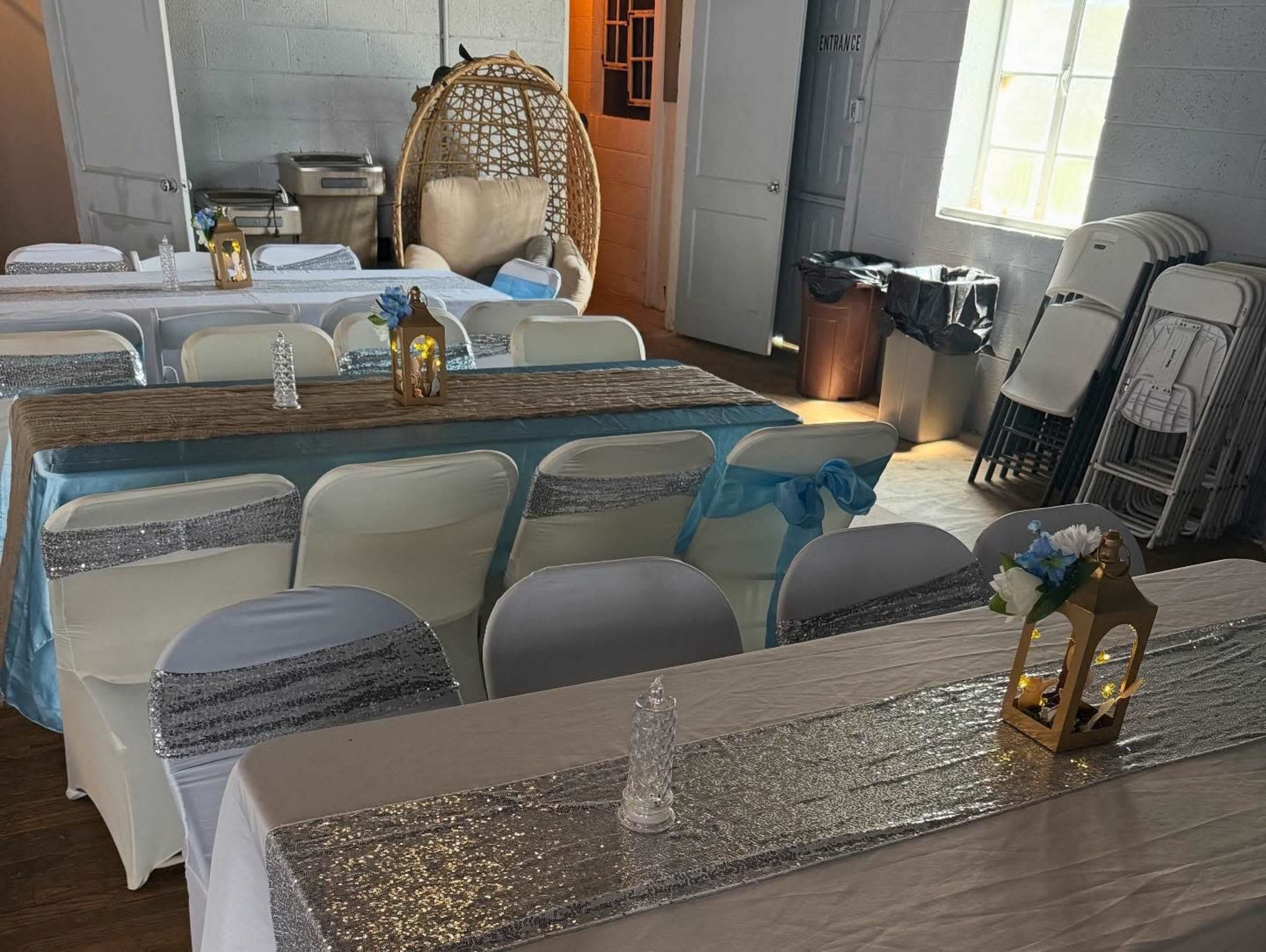 The image shows a decorated indoor space set up for an event, featuring tables with white tablecloths and centerpieces, alongside chairs draped in white covers with blue accents.