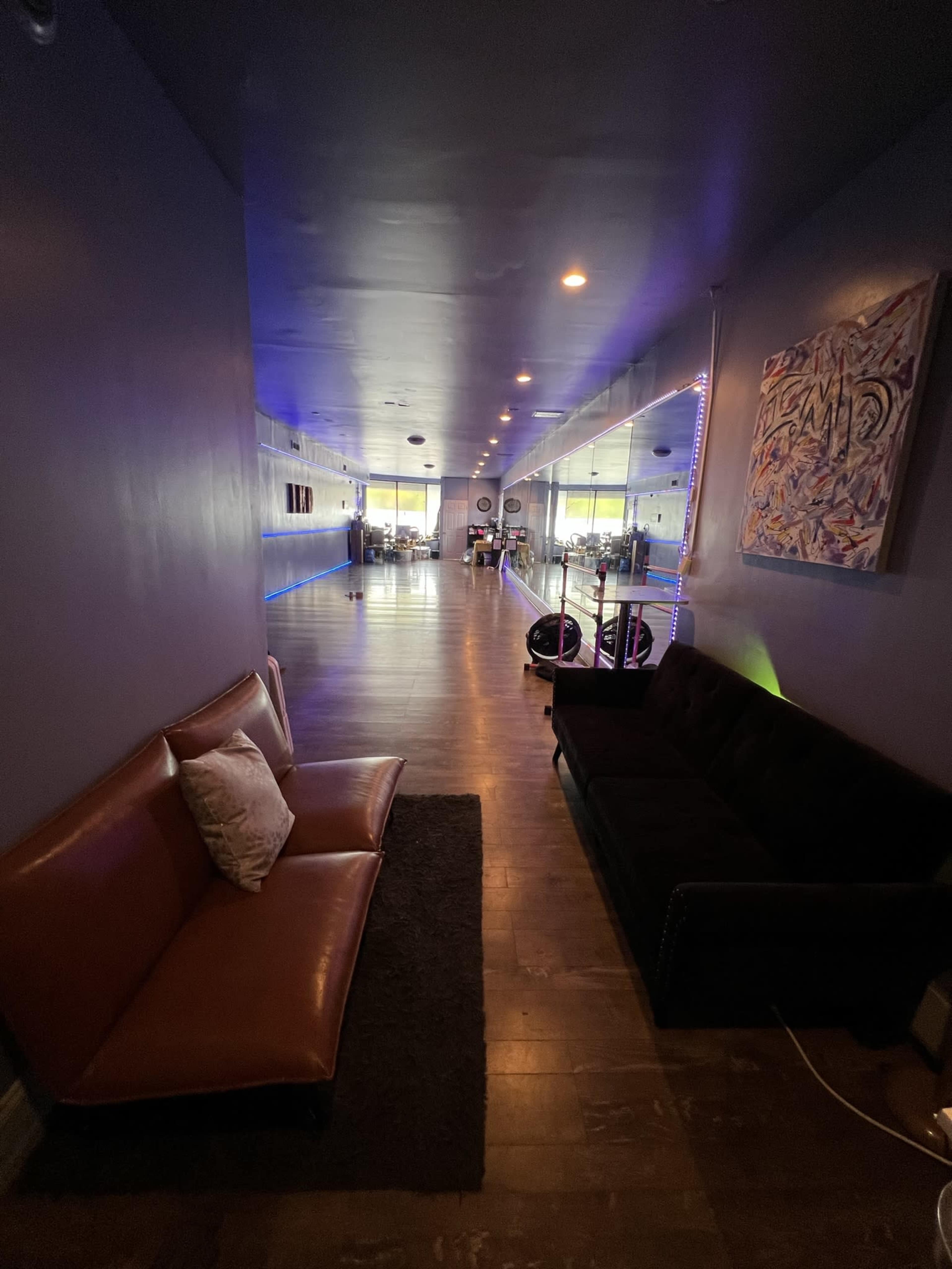 A hallway with two sofas along the side, illuminated by blue lighting, leading towards a spacious area with various furnishings in the background.