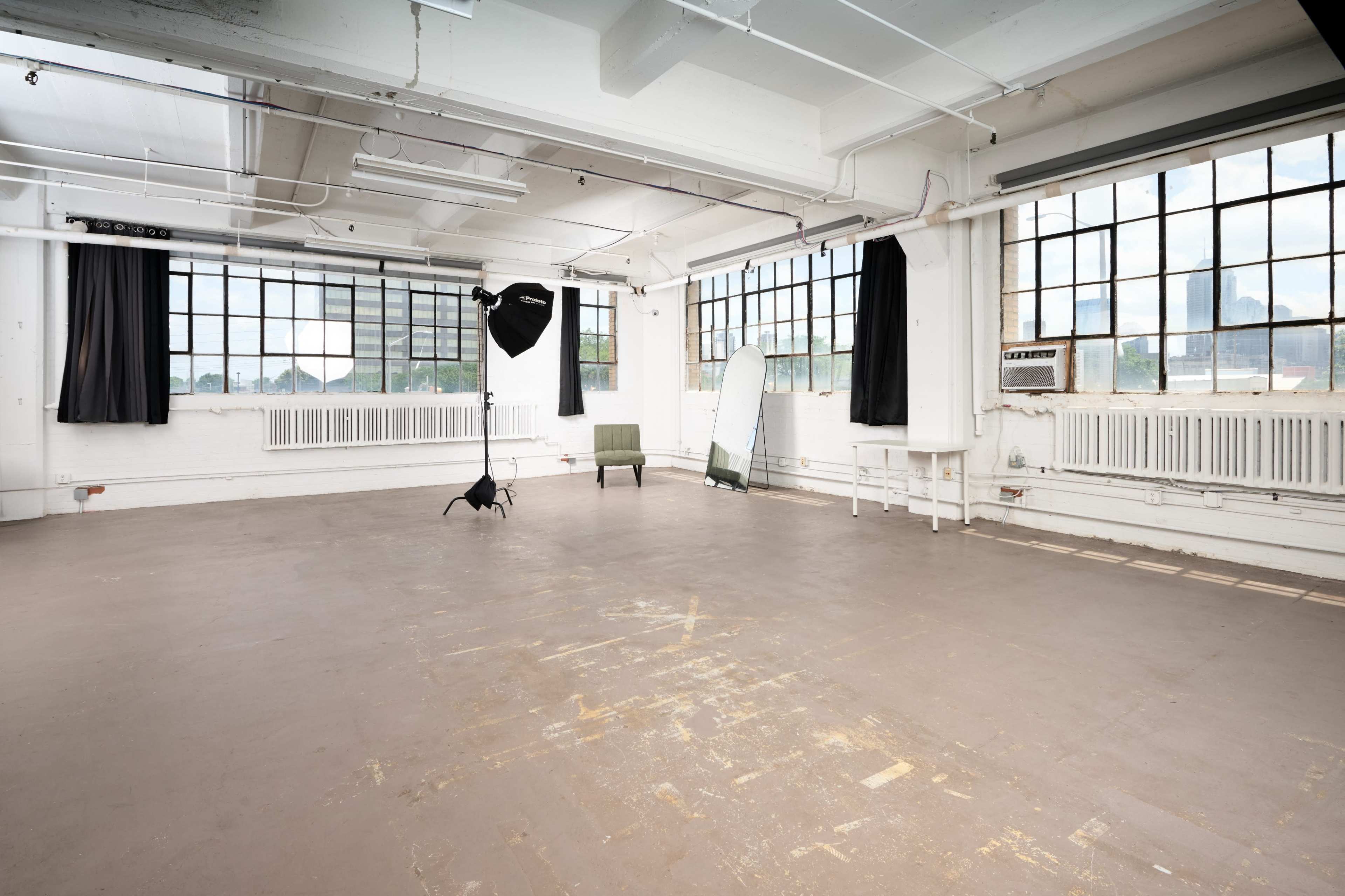 The image shows a spacious, empty studio with large windows, a green chair, a mirror, and photography equipment set up in one corner.