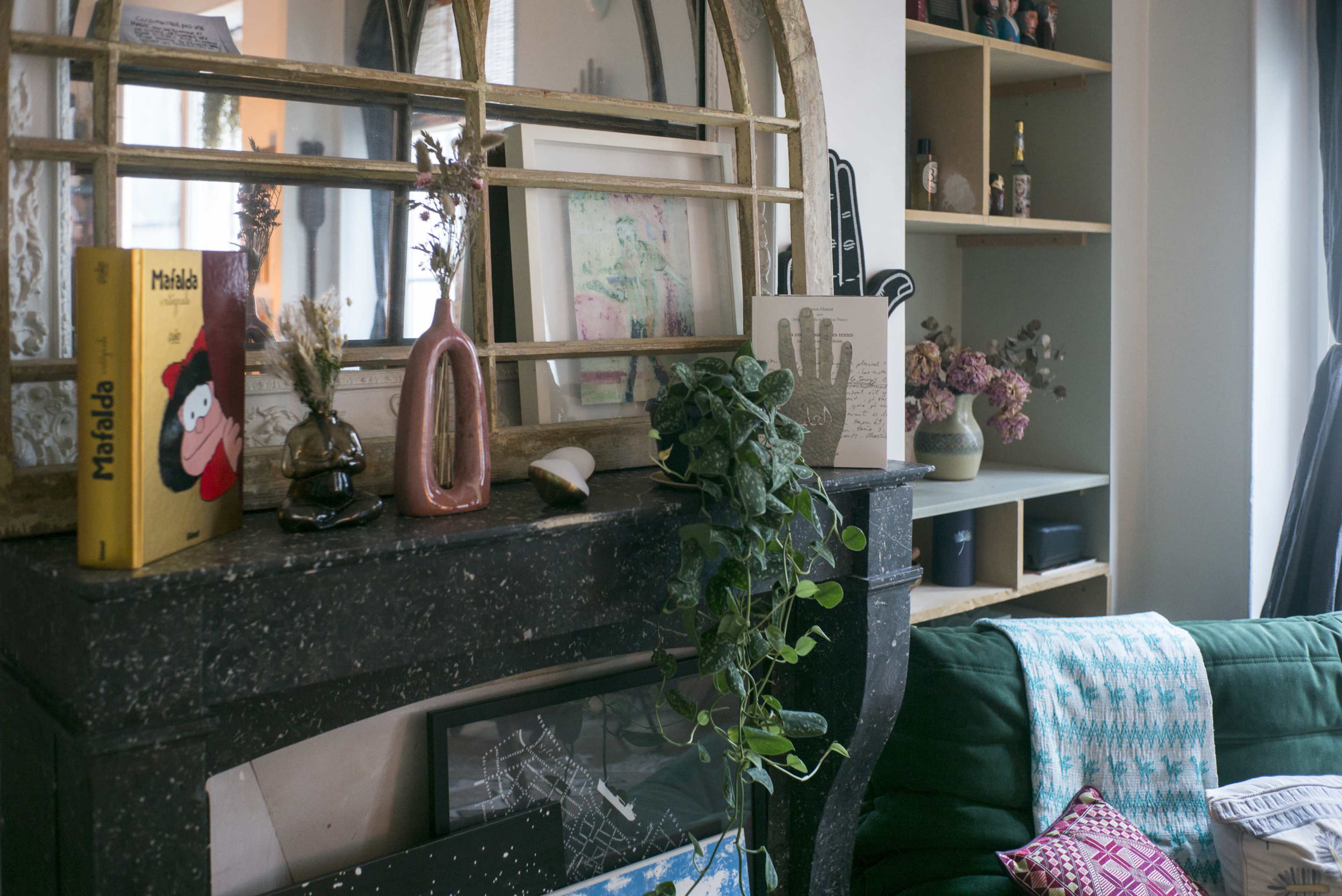 A decorative living room features a stone fireplace mantel adorned with books, vases, and plants, with a large mirrored arch above it.
