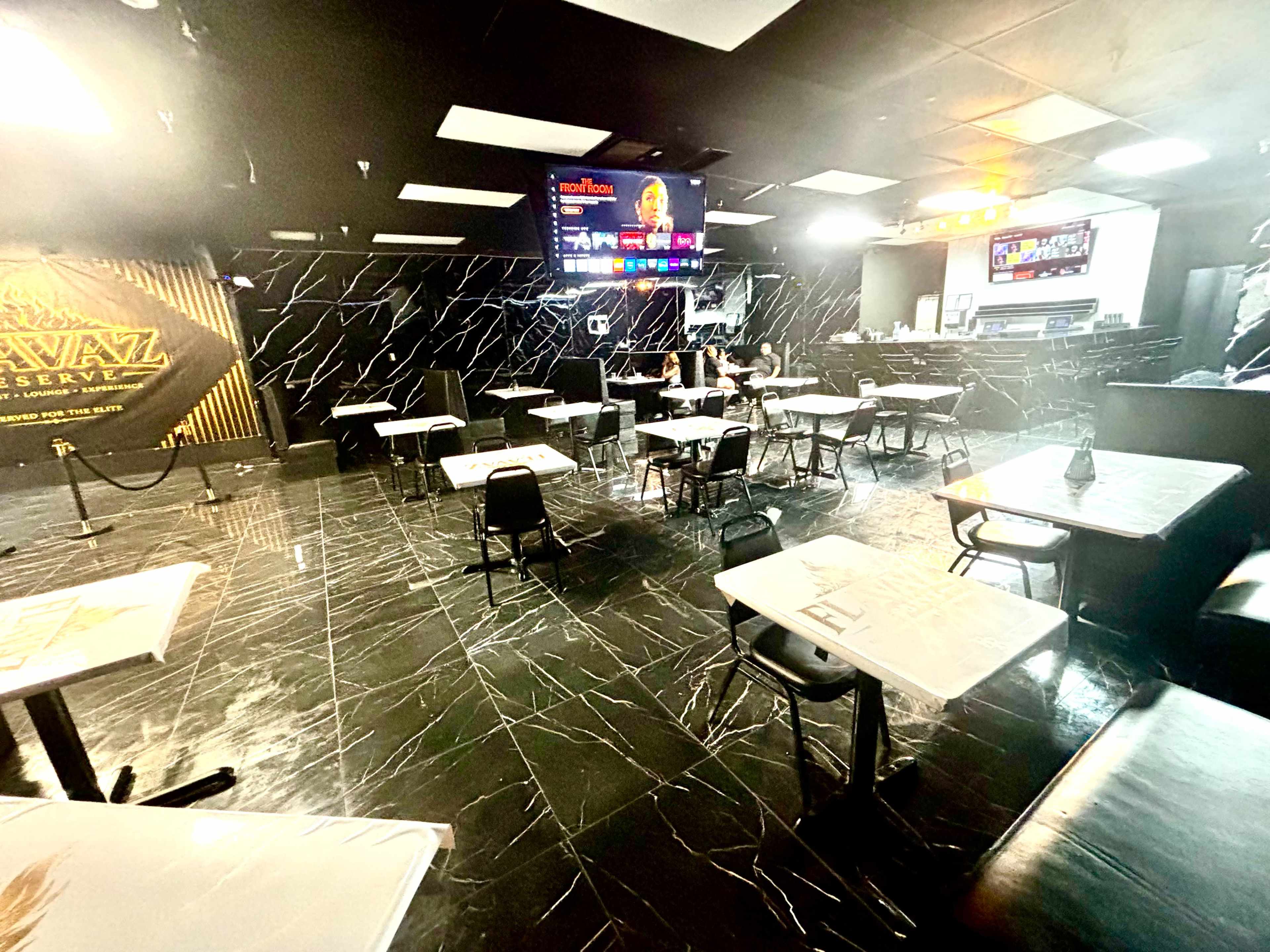 The image shows a modern restaurant interior with black marble floors, multiple tables and chairs, and a television displaying news in the background.