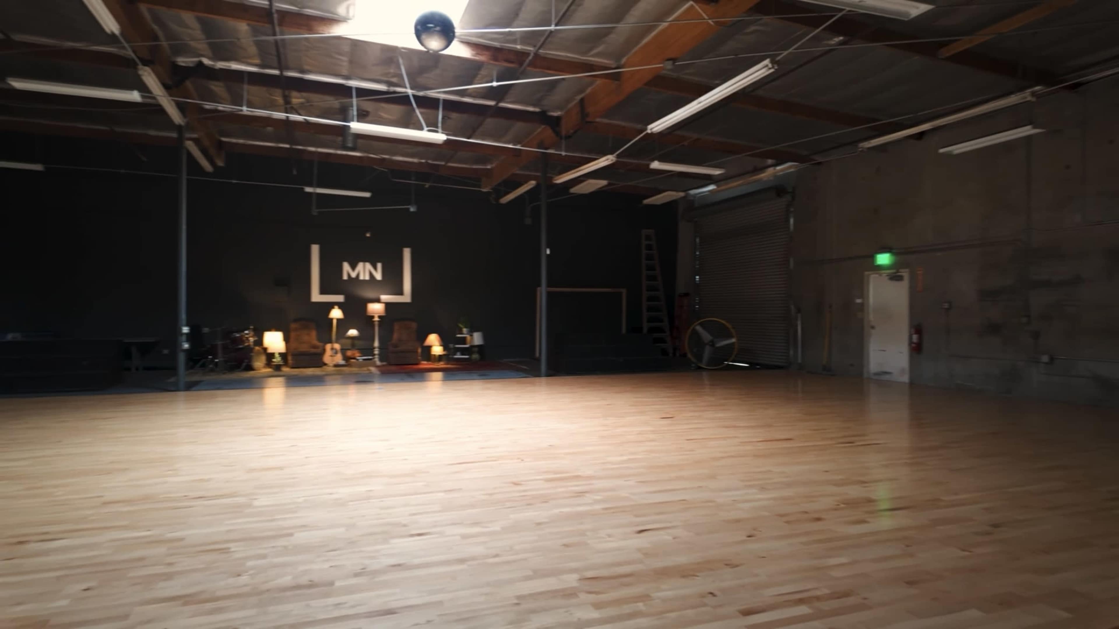 A spacious dance studio with wooden flooring, high ceilings, and minimal furnishings, illuminated by overhead lights.