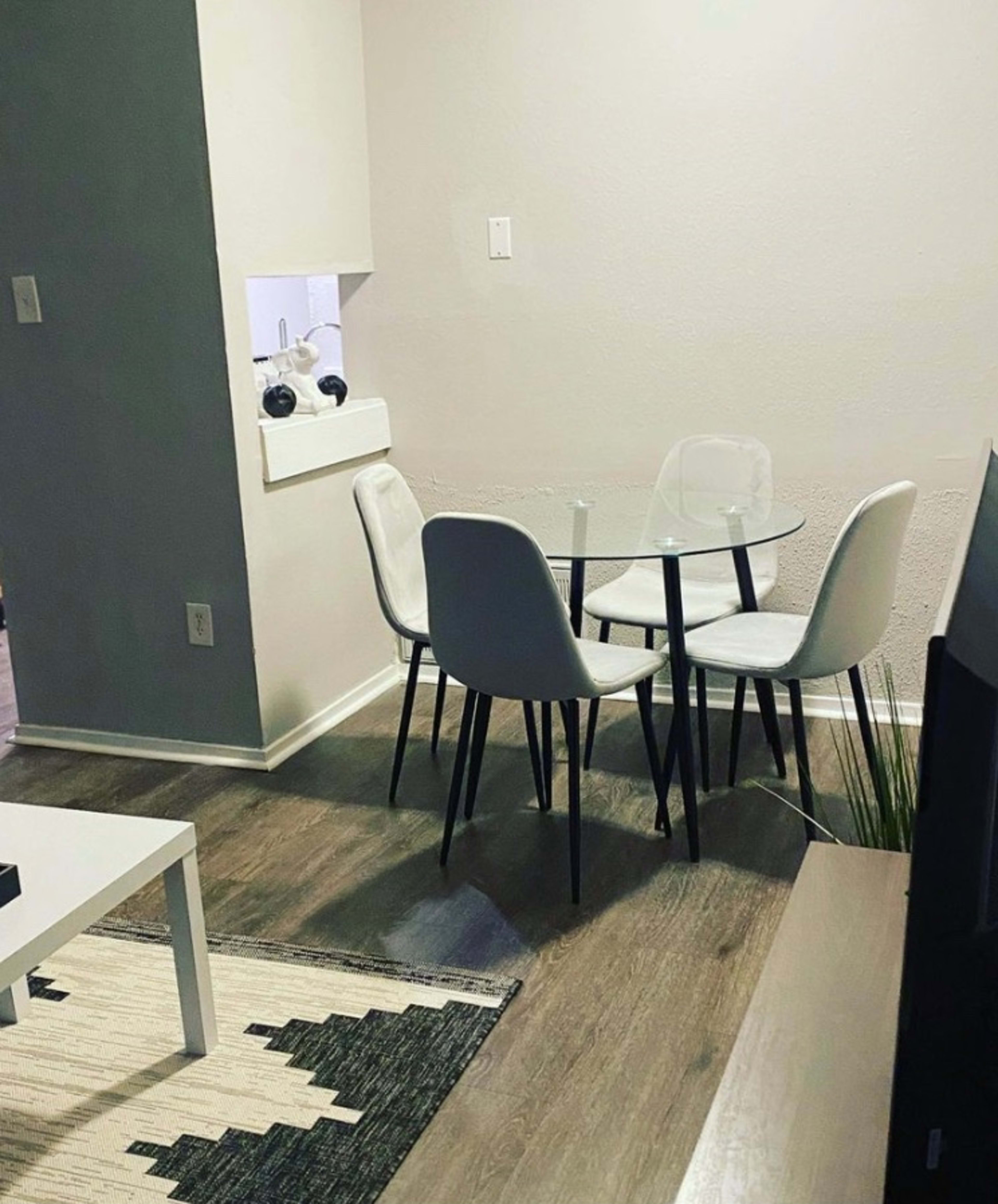 A small dining area features a glass table surrounded by four white chairs, with a view of a decorative rug and a plant nearby.