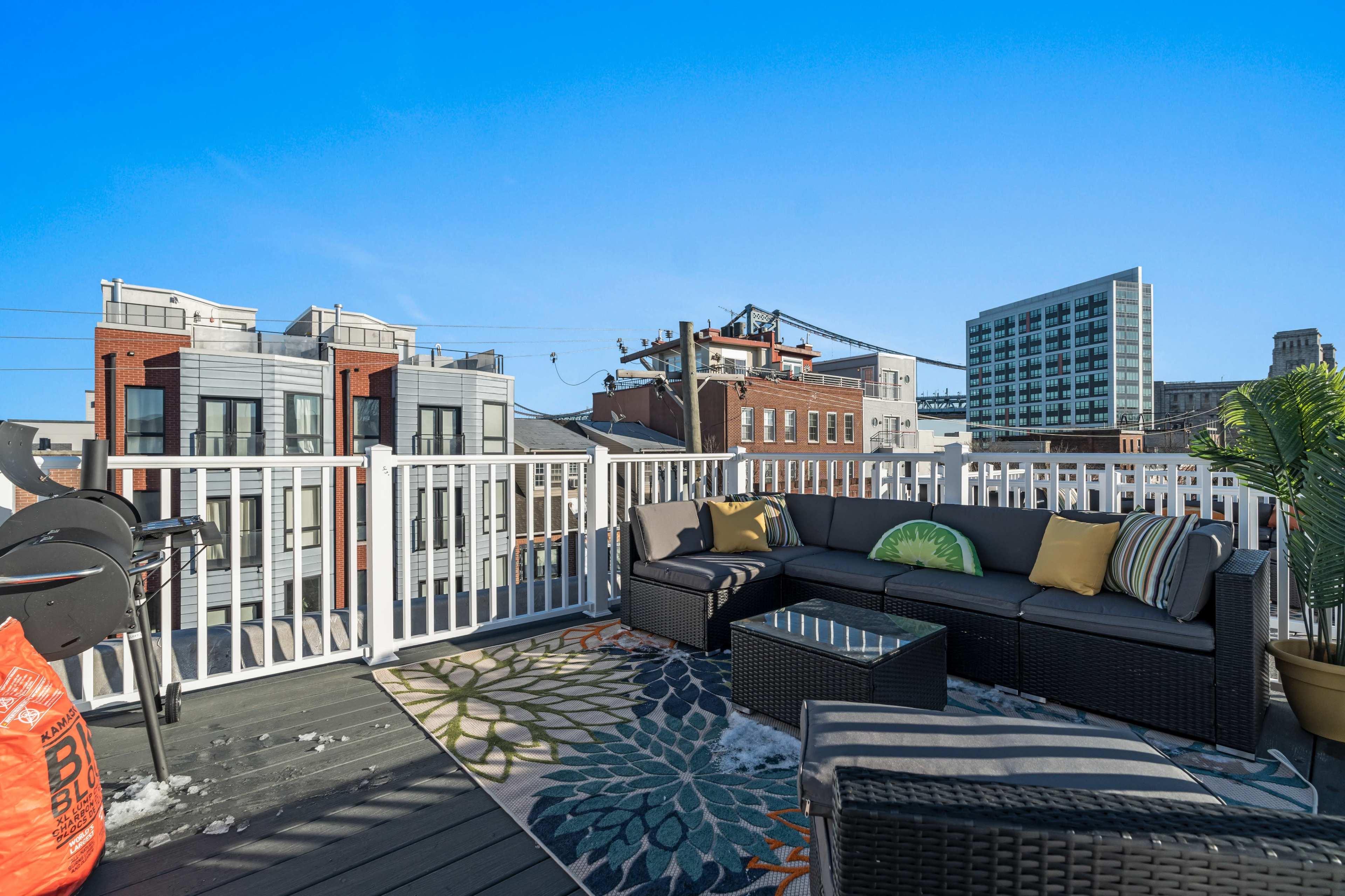 The image shows a rooftop terrace furnished with a large sectional sofa, a coffee table, and decorative plants, overlooking a cityscape with various buildings and a clear blue sky.