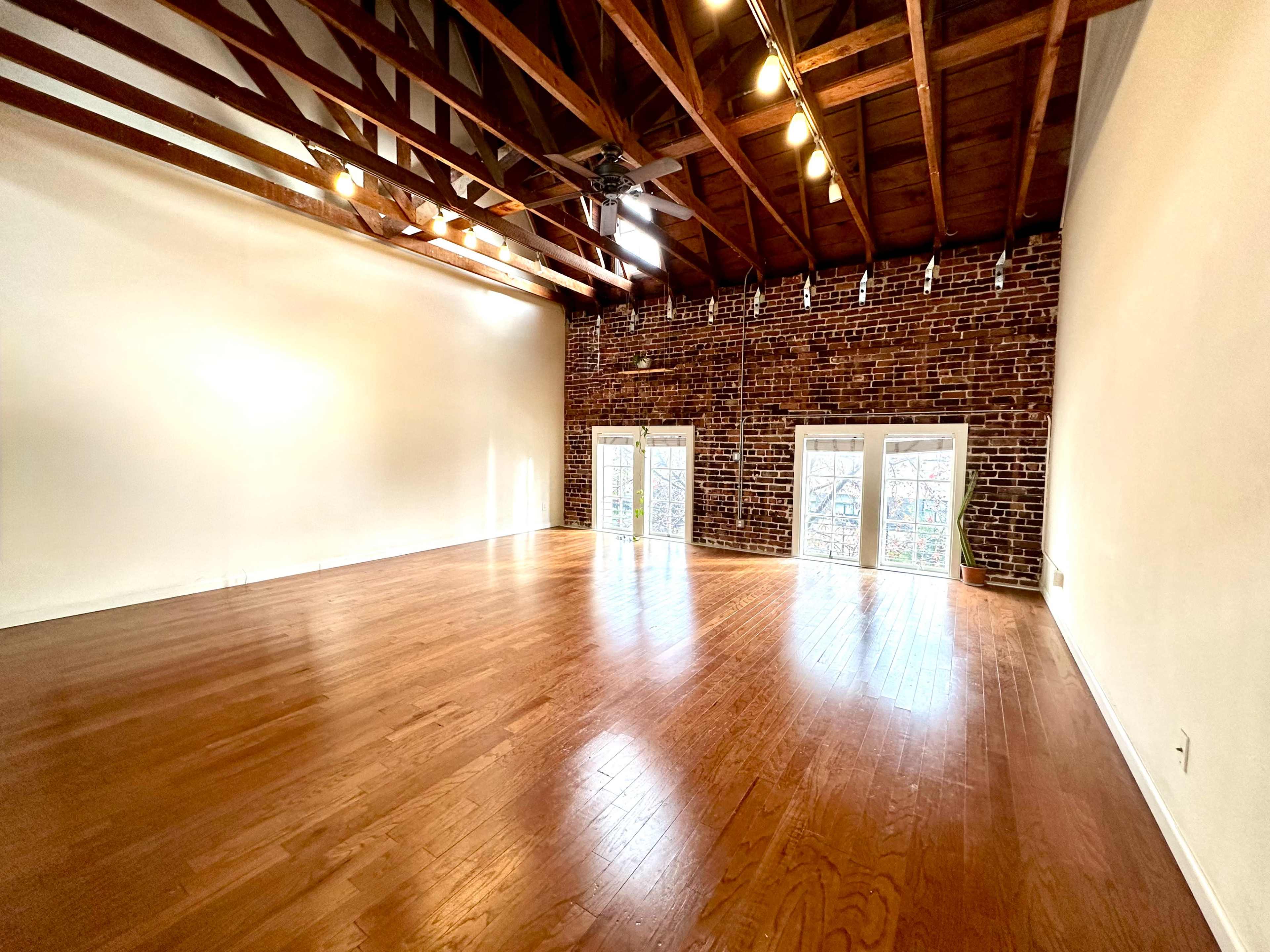 The image shows an empty room with wooden floors, exposed brick walls, and beam ceilings, illuminated by natural light from large windows.