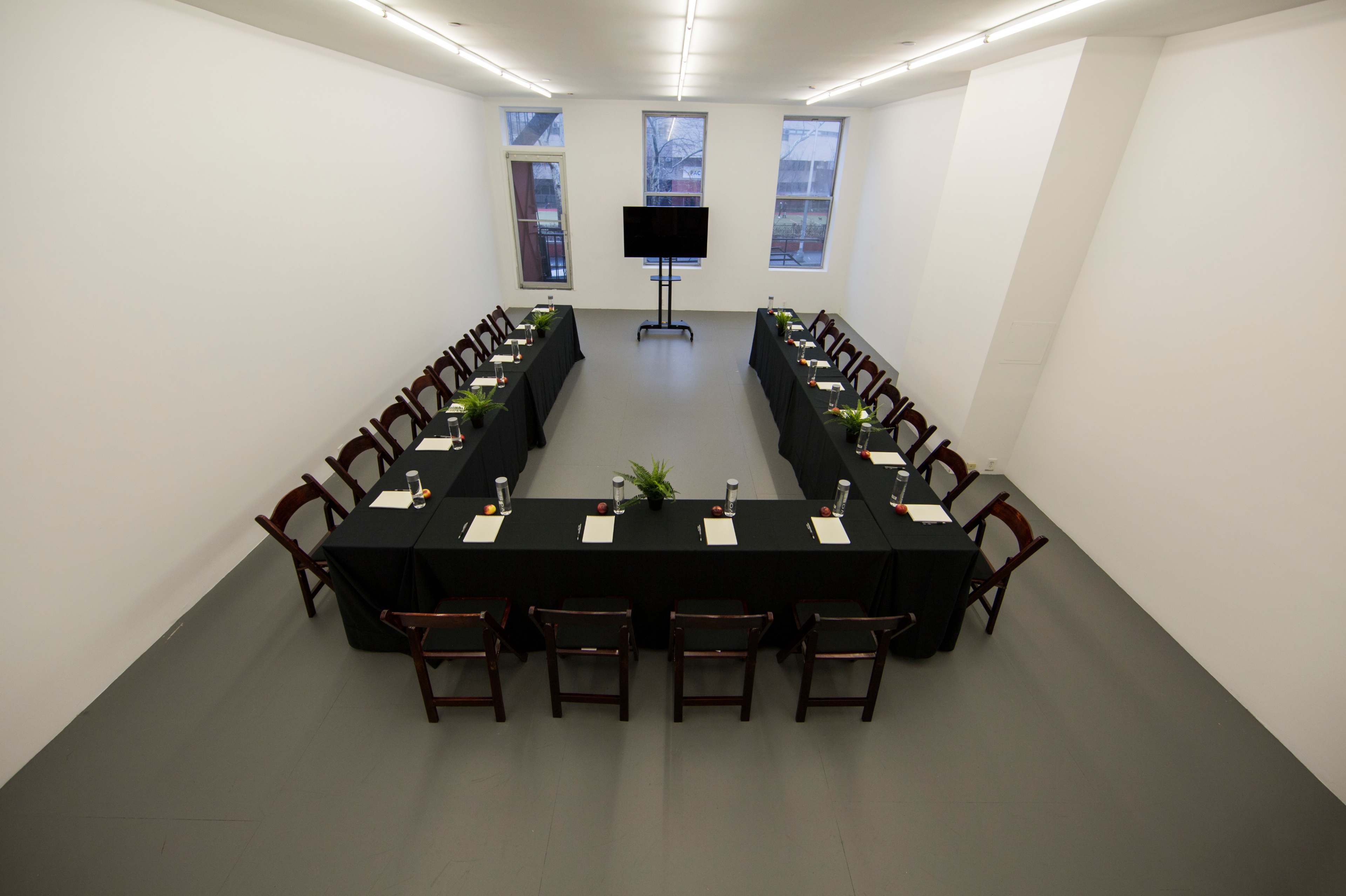 A rectangular conference room features a long table set with tableware and surrounded by wooden chairs, with a television screen at one end and large windows on the walls.