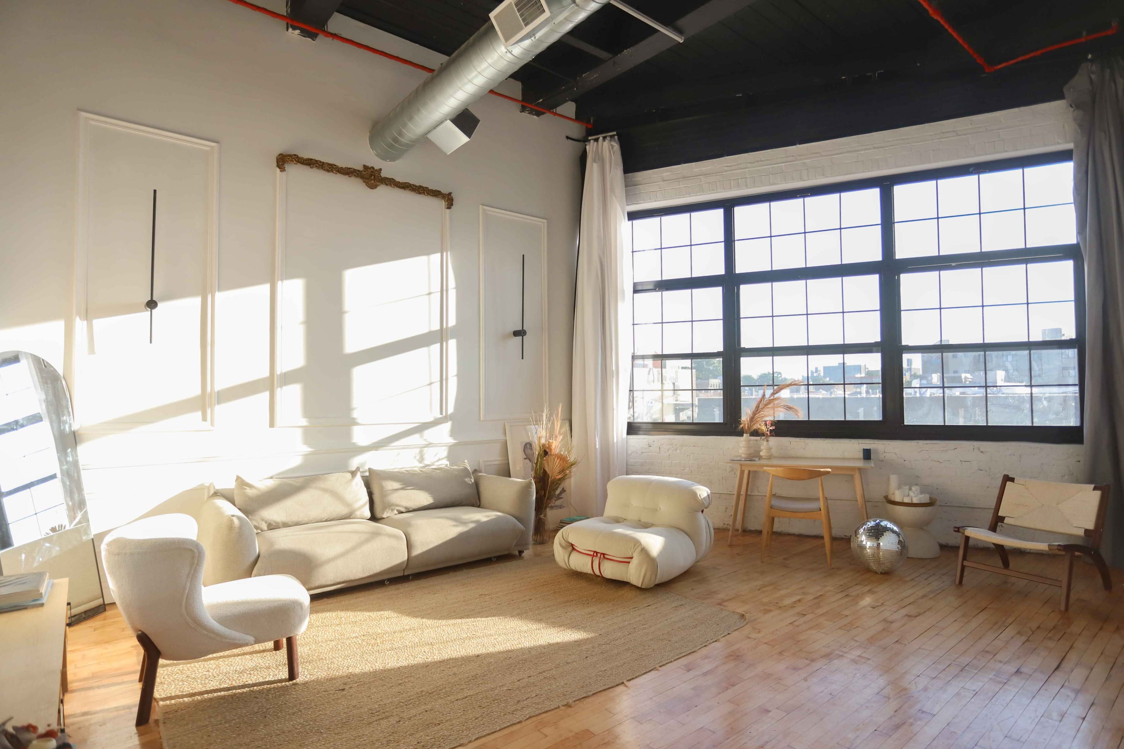 A well-lit interior space features a light-colored sofa, a large window, and minimalist decor, with wooden flooring and a woven rug.
