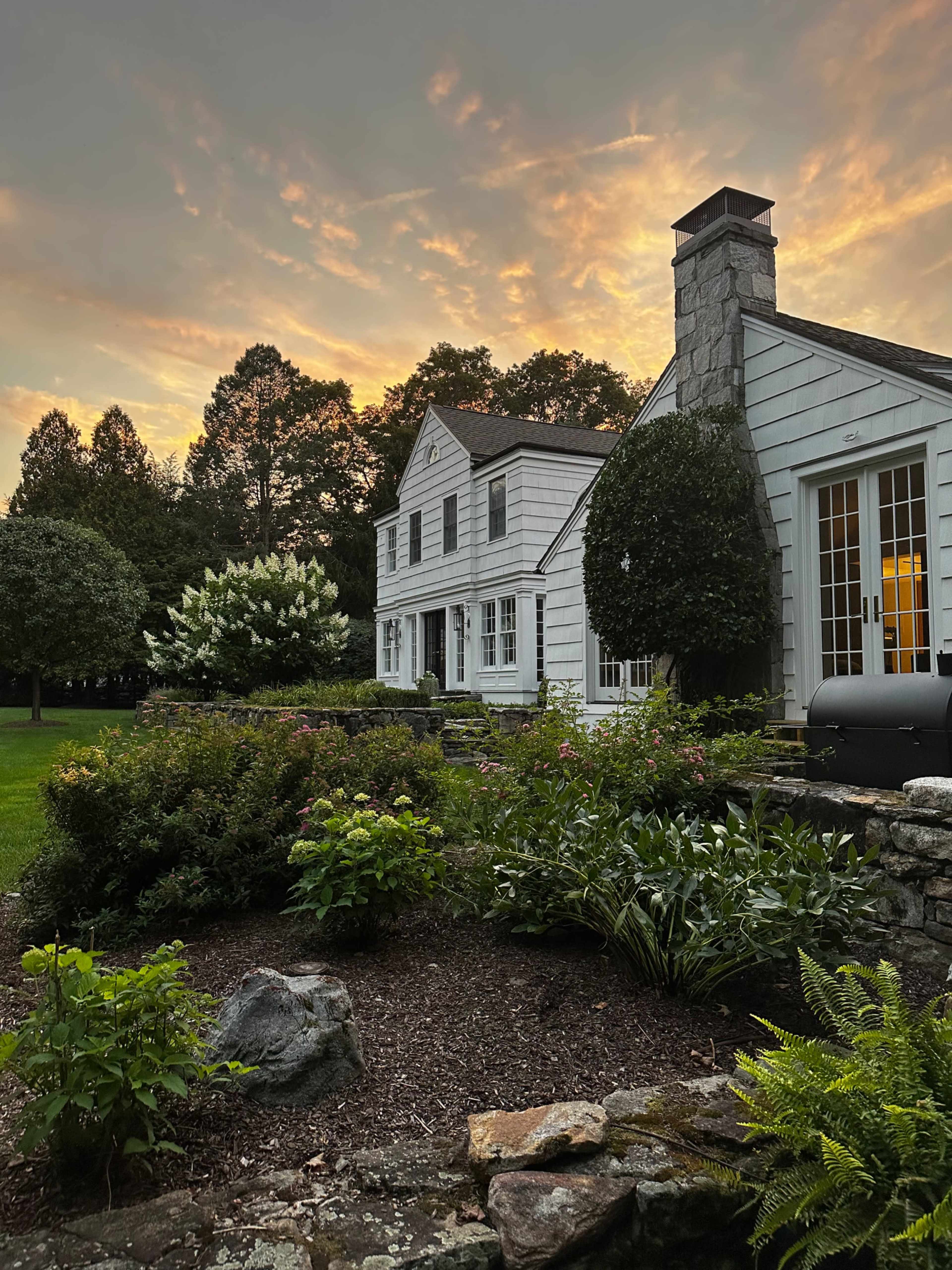 A white two-story house with a stone chimney is surrounded by well-maintained gardens under a colorful sunset sky.