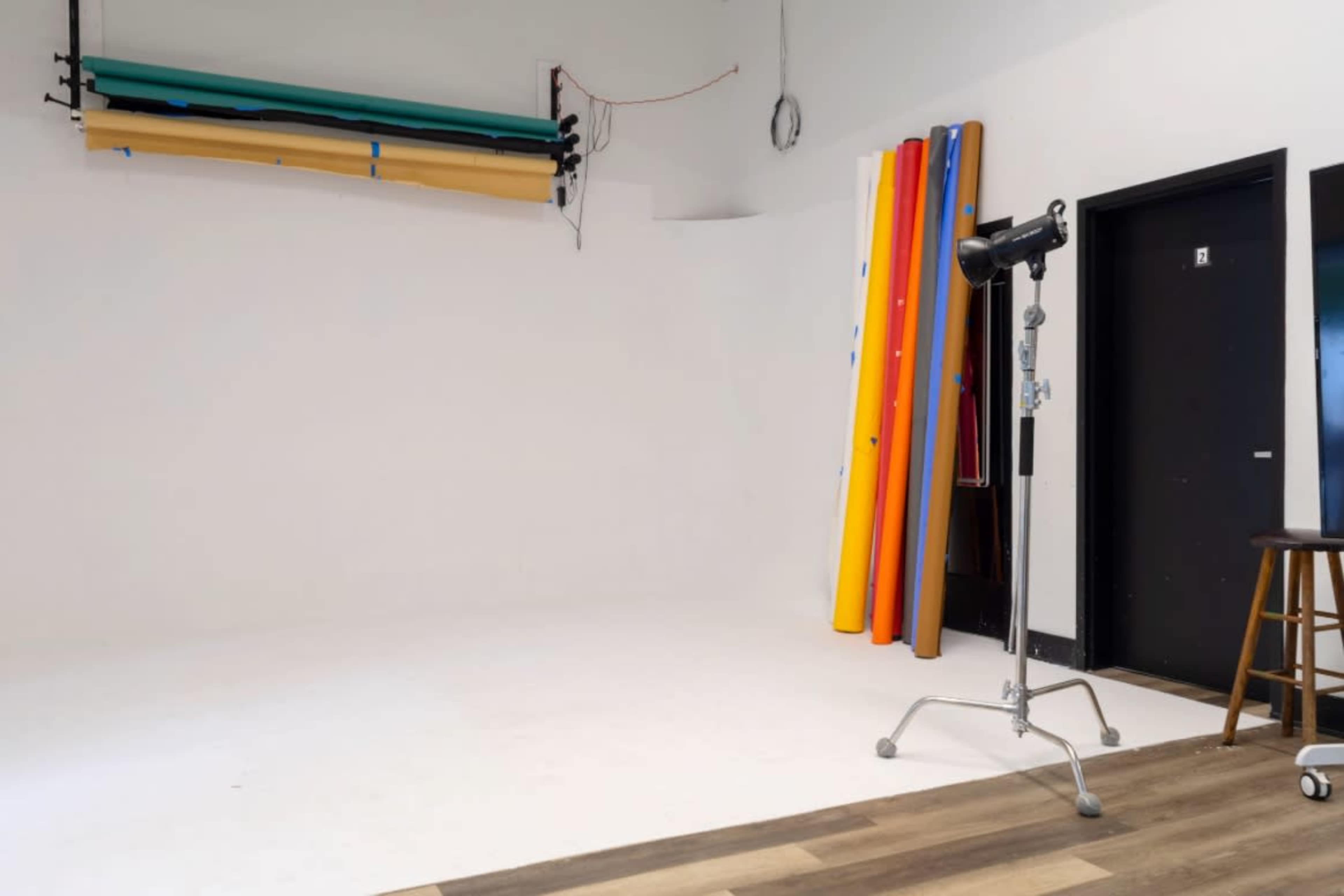The image shows a photography studio with a blank white backdrop, a standing light fixture, a wooden stool, and colorful backdrops leaning against the wall.