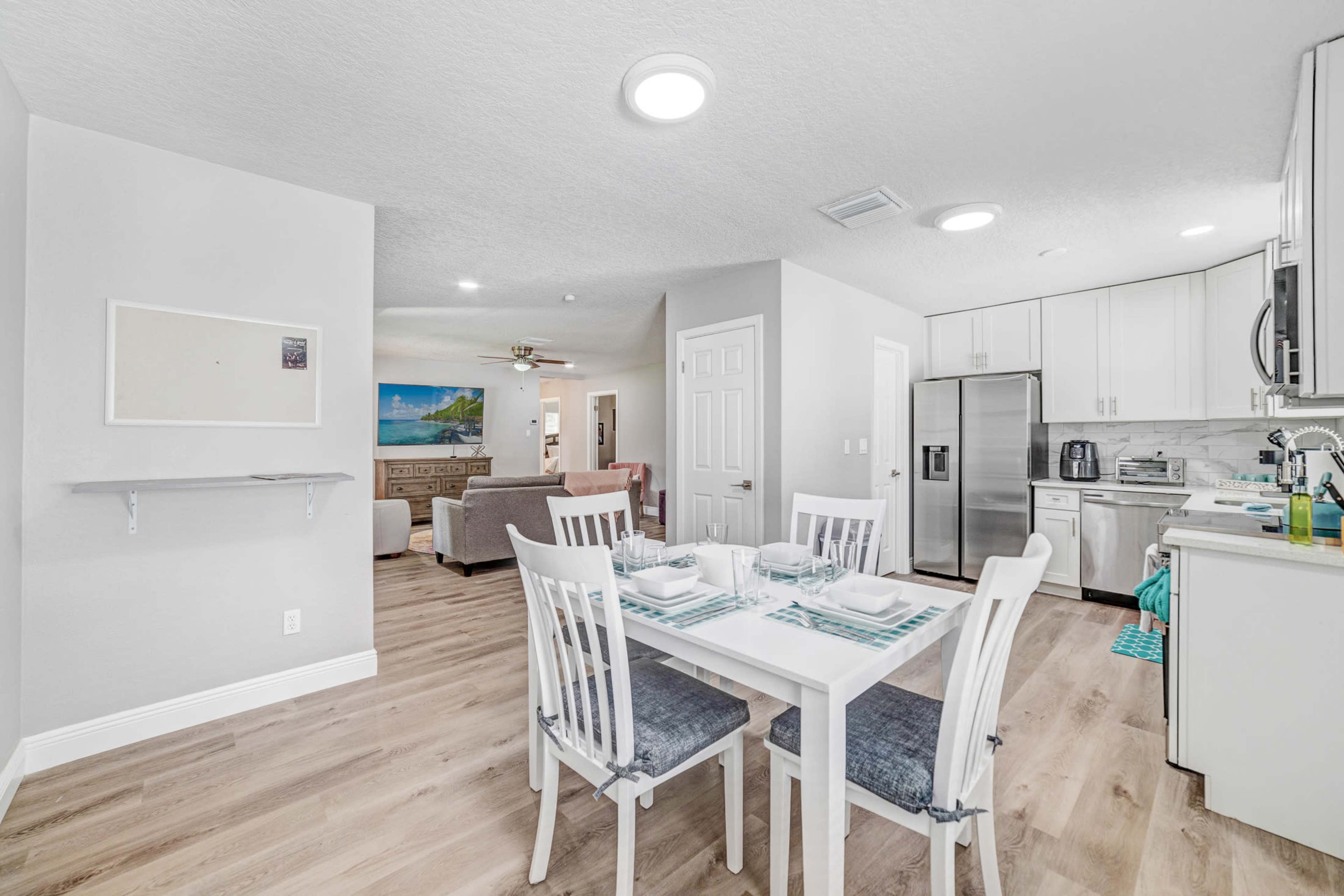 A modern, open-concept kitchen and dining area with a white table set for four, light-colored cabinetry, and a cozy living room visible in the background.