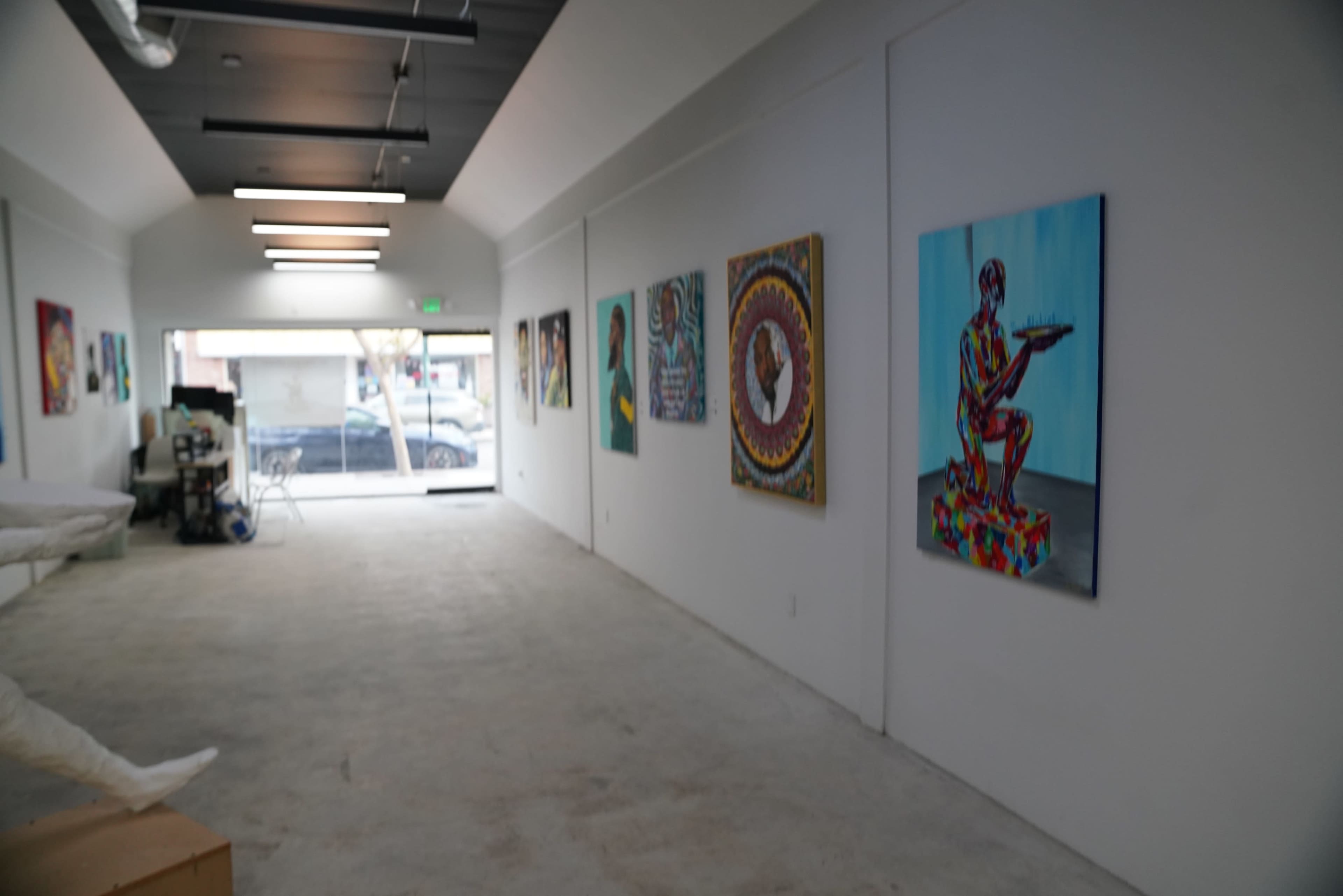 The image shows a gallery-like interior with colorful artwork displayed on white walls and large windows at the far end.