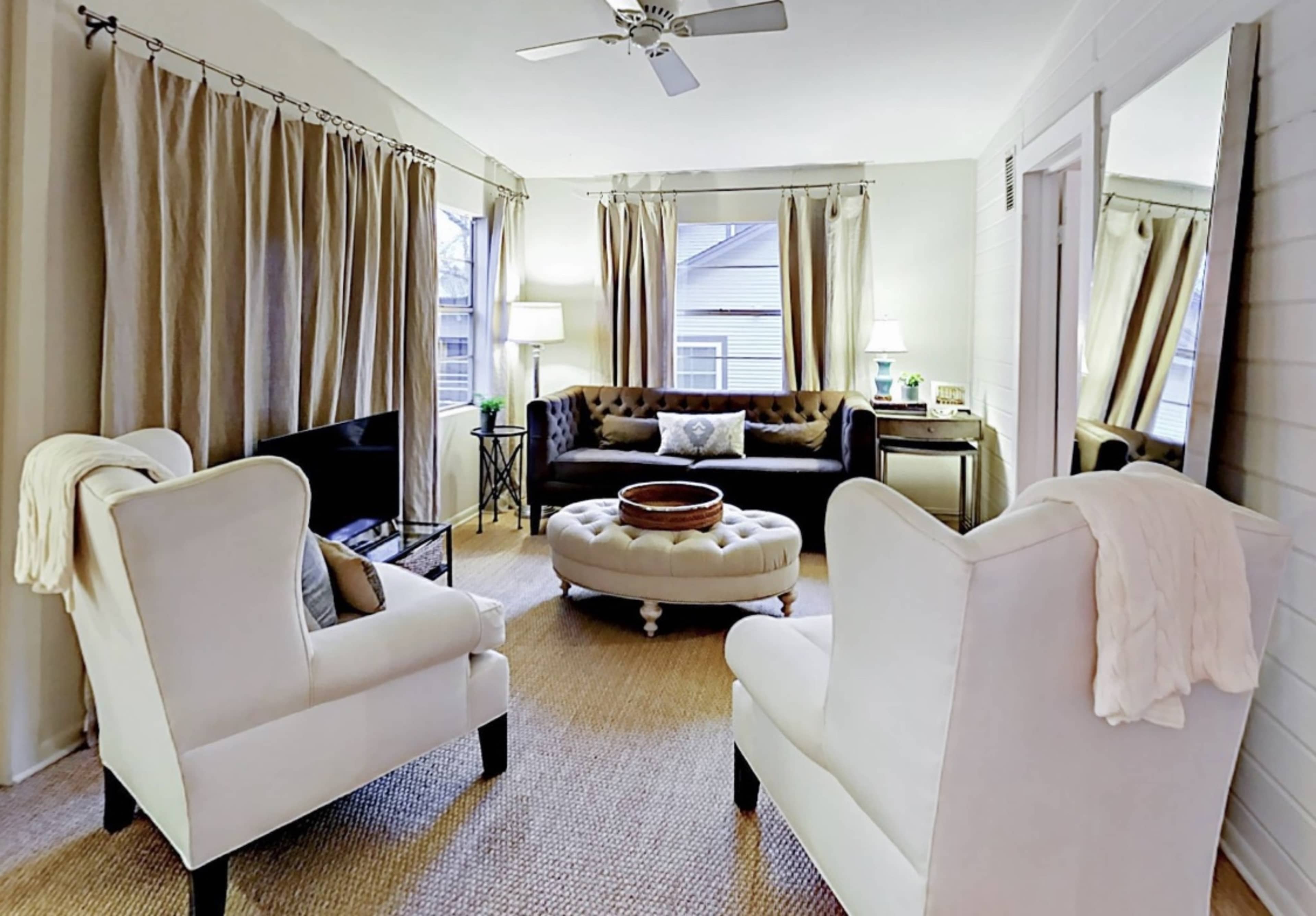 A cozy living room with a tufted sofa, two white armchairs, a round ottoman, and large windows covered with beige curtains.