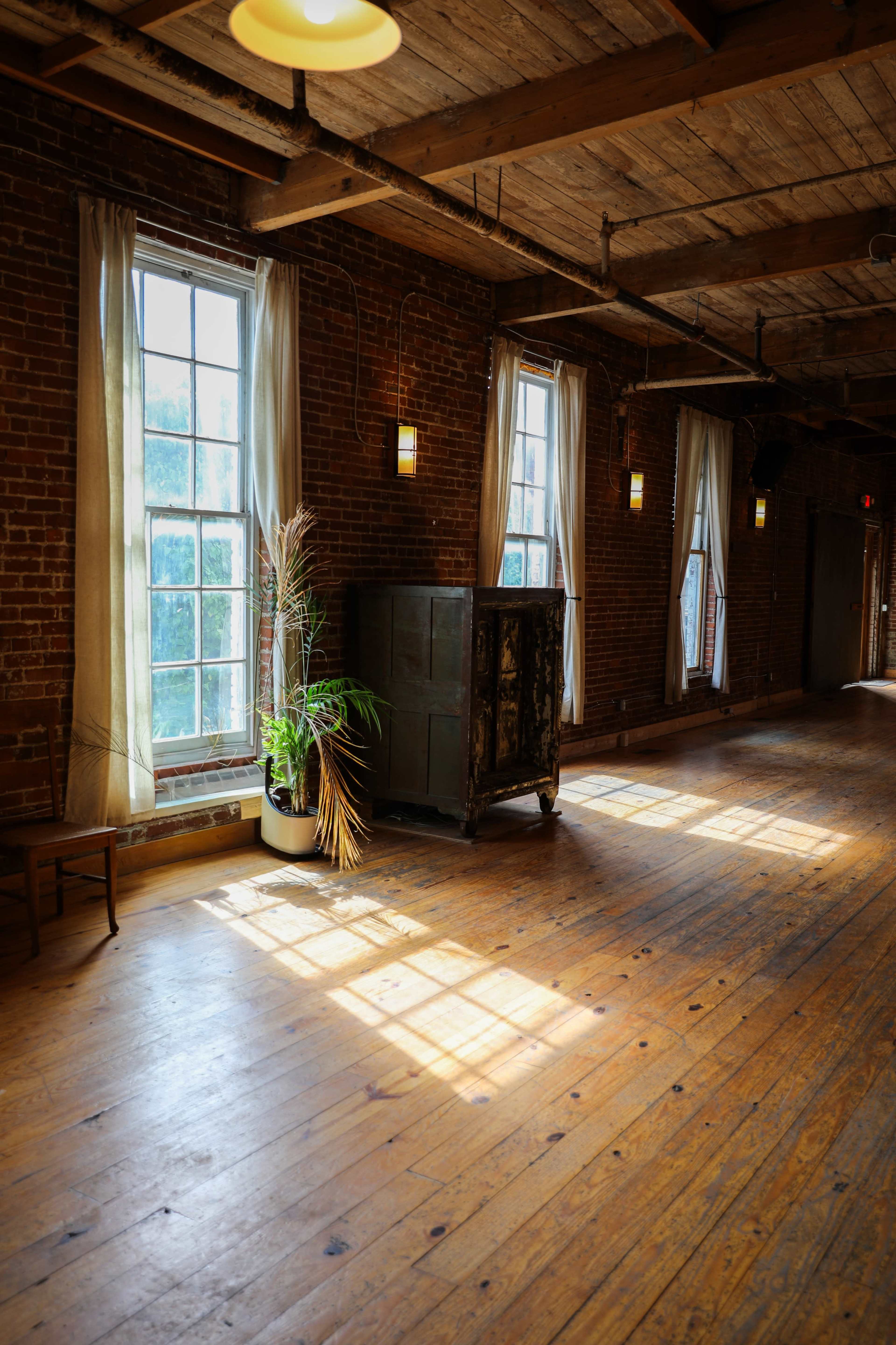 A rustic room with wooden floors features large windows, a green plant next to an old wooden cabinet, and soft natural light streaming through the space.