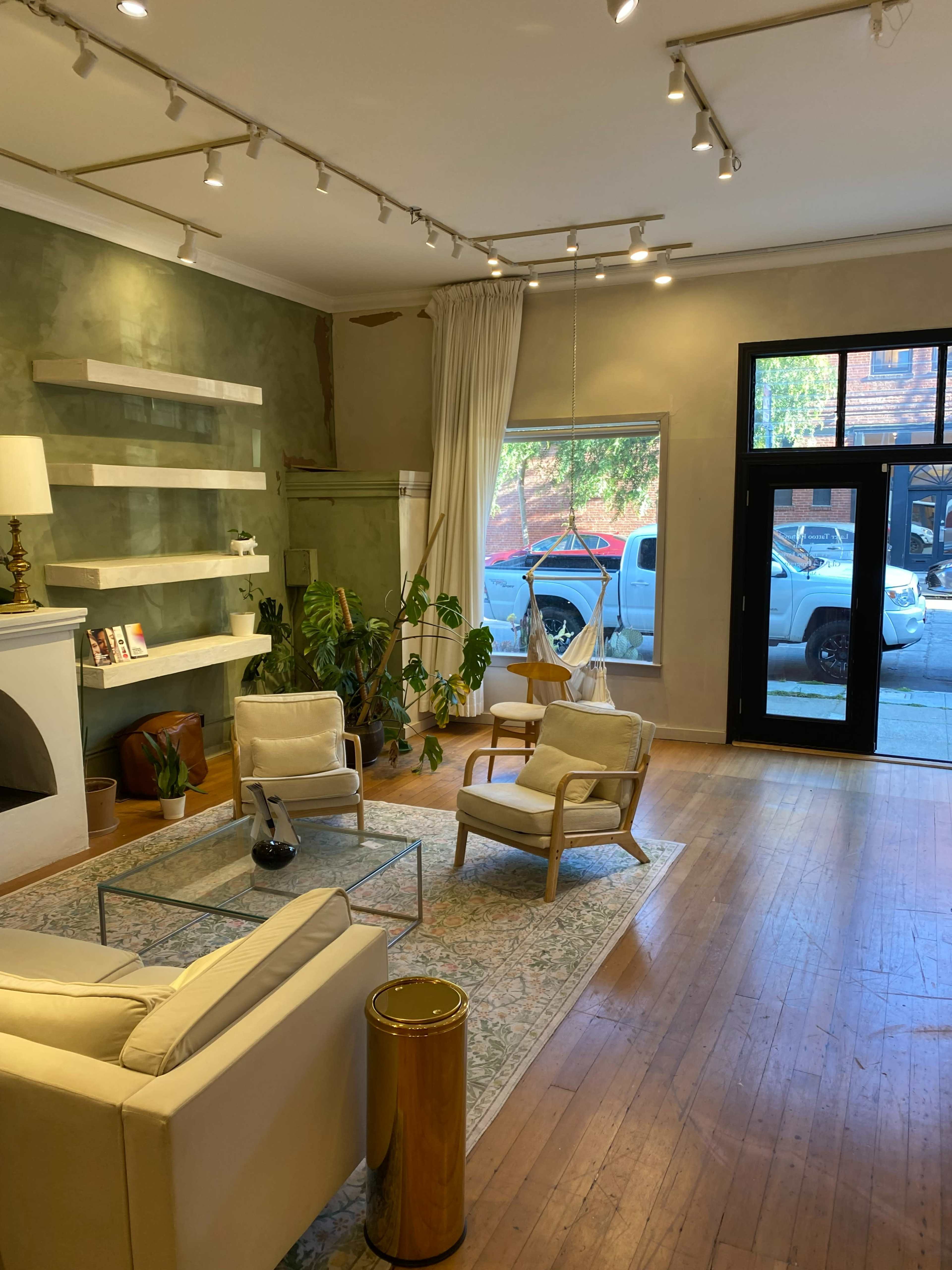 The image shows a well-lit interior of a modern living space with two armchairs, a sofa, a glass coffee table, and a large window facing the street.