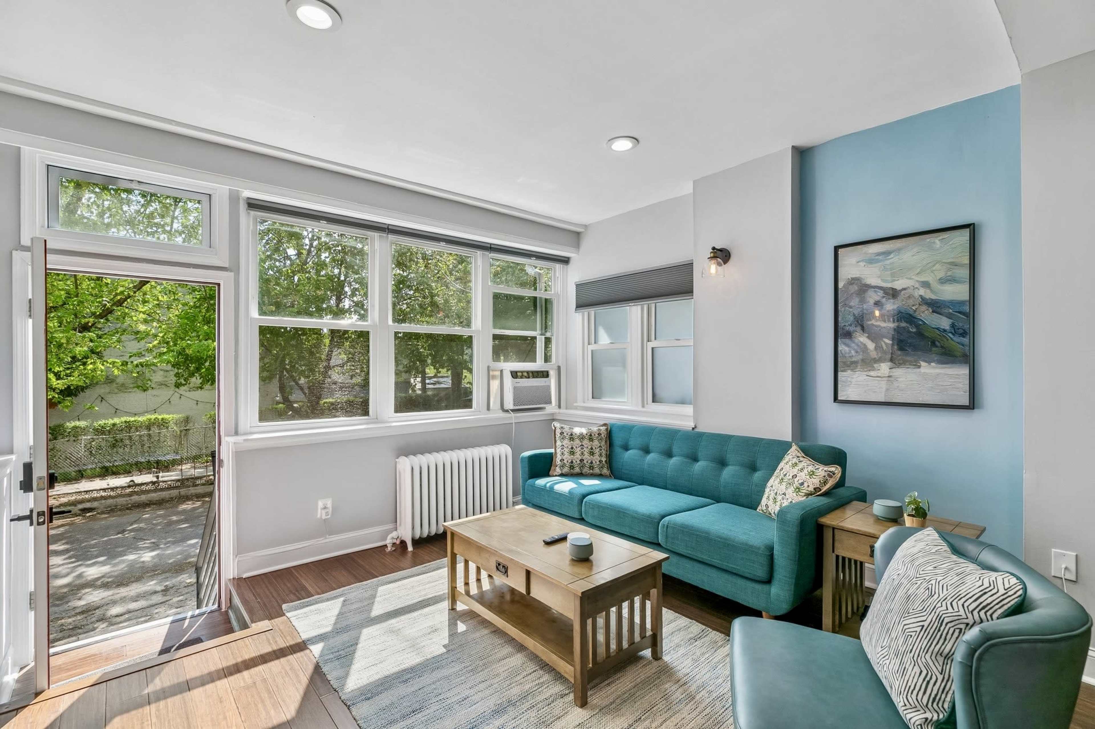 The image shows a sunlit living room with a turquoise couch, a wooden coffee table, and large windows overlooking greenery outside.