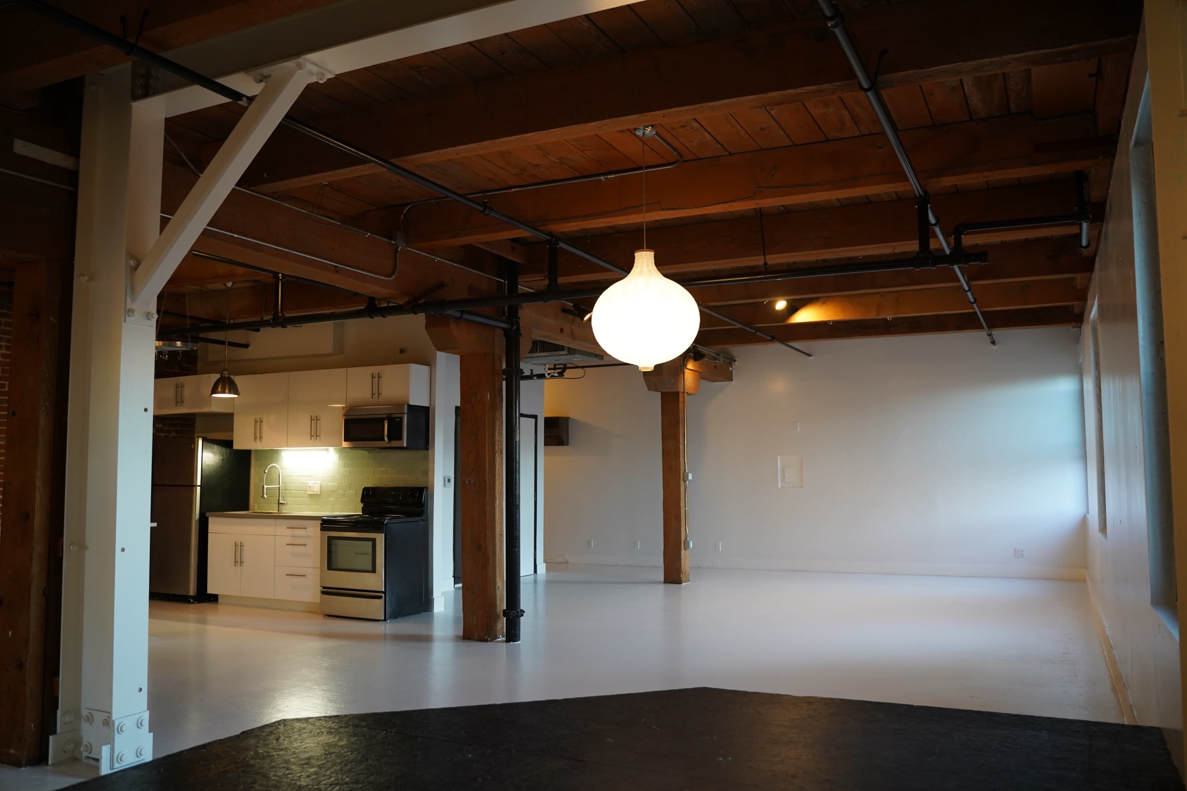 The image shows a spacious, open-concept room with wooden beams, a simple kitchen area, and a large pendant light hanging from the ceiling.