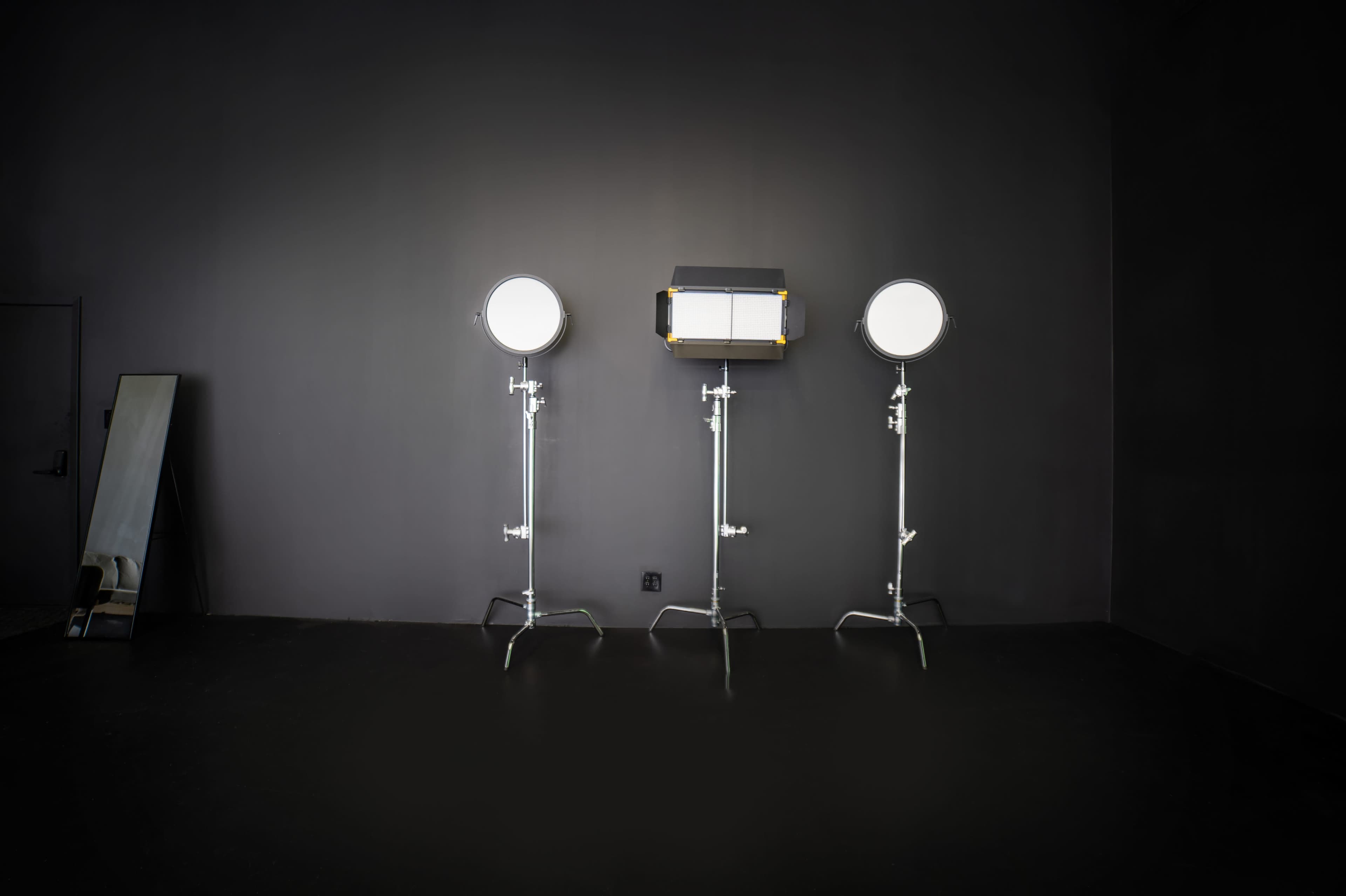 Three studio lights positioned on stands in a dark room with a mirror leaning against the wall.