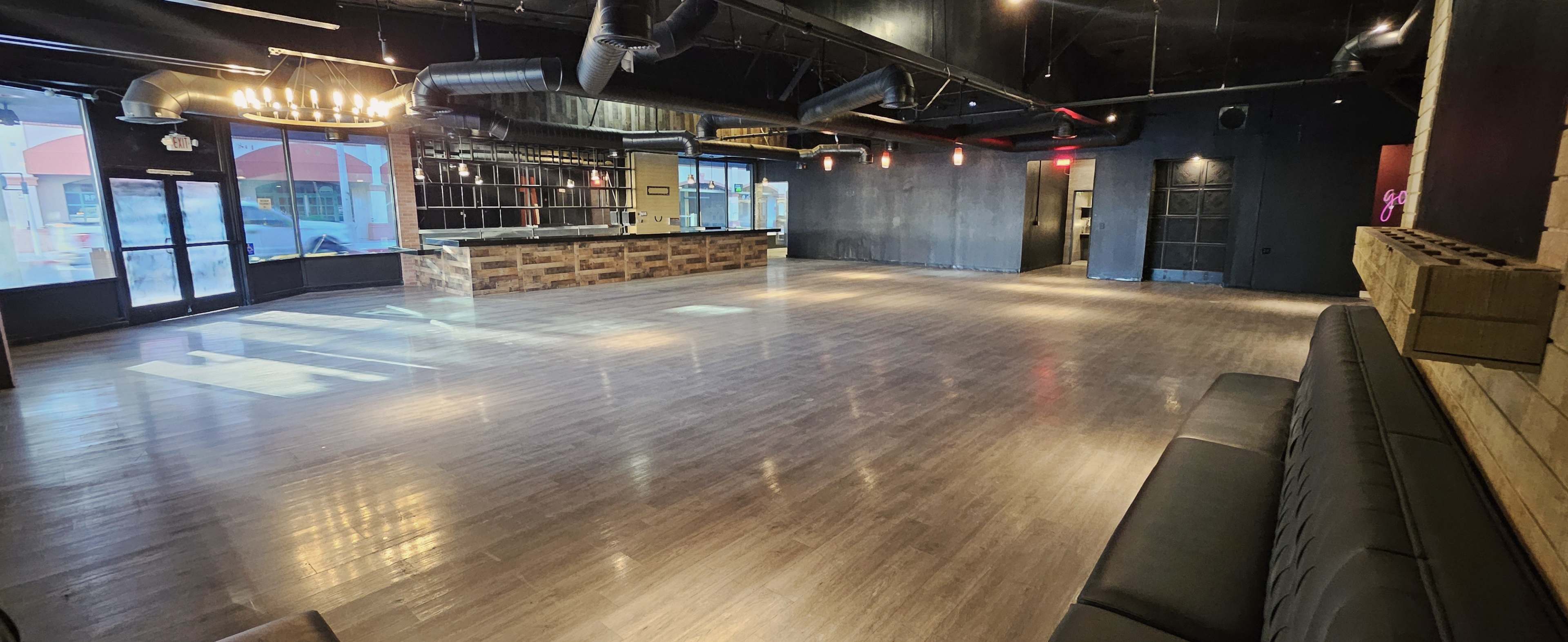The image shows a spacious, empty venue with a polished wooden floor, large windows, and a bar area in the background against a dark-colored interior.