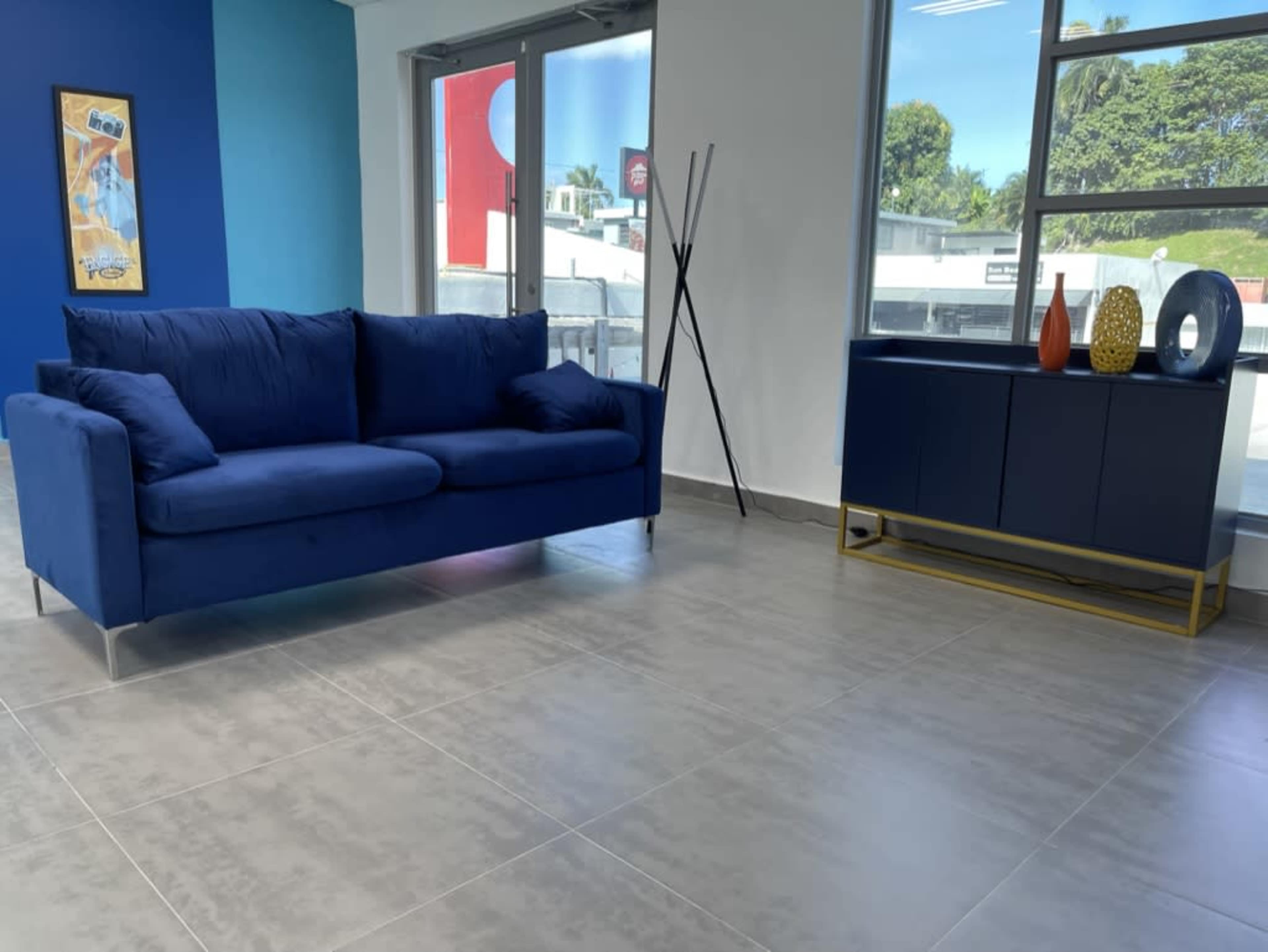 The image shows a modern interior featuring a blue couch, a dark cabinet with decorative items, and large windows with natural light.