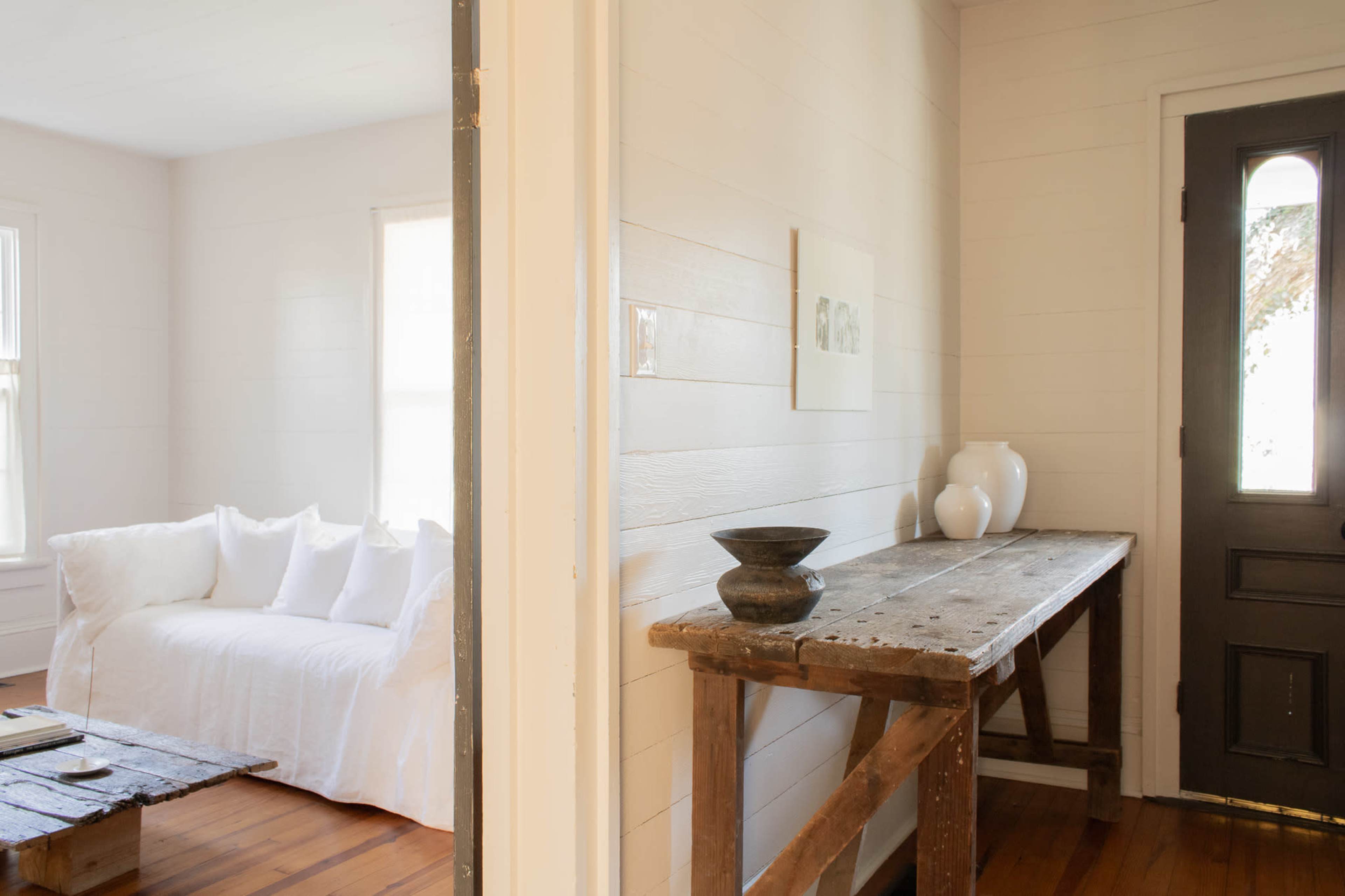 The image shows a bright, minimalistic interior with a white sofa and a wooden table near an entrance door.