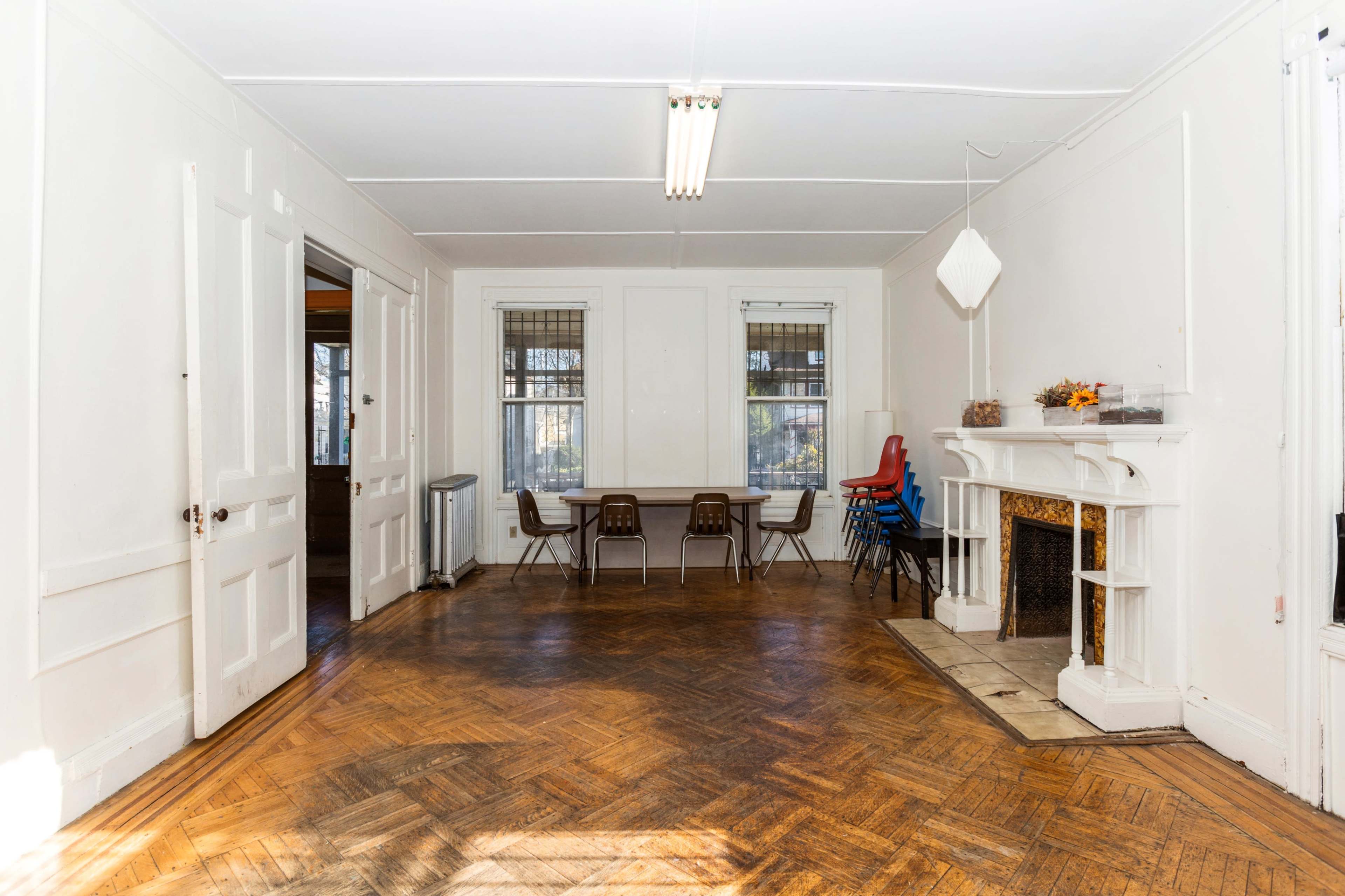 The image shows a large, empty room with a herringbone wooden floor, windows on two walls, and a fireplace with a decorative mantel.